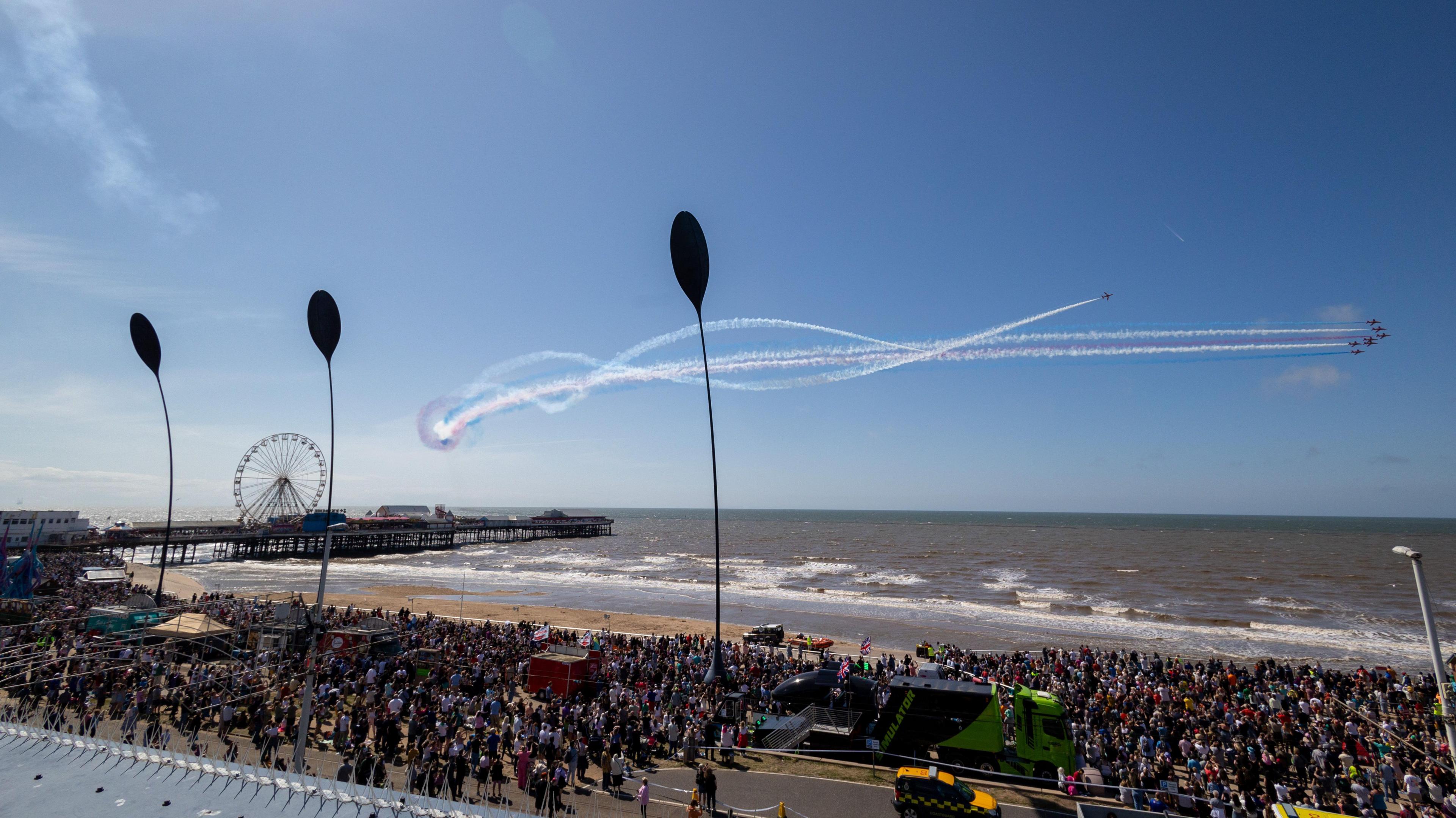 A photo of a sunny day at Blackpool beach. thousands of people can be seen in the crowd on the seafront. In the blue skies above, planes are flying from left to right, leaving trails of red white and blue. A pier with a wheel can be seen in the distance, and there are a few stalls on the beach.