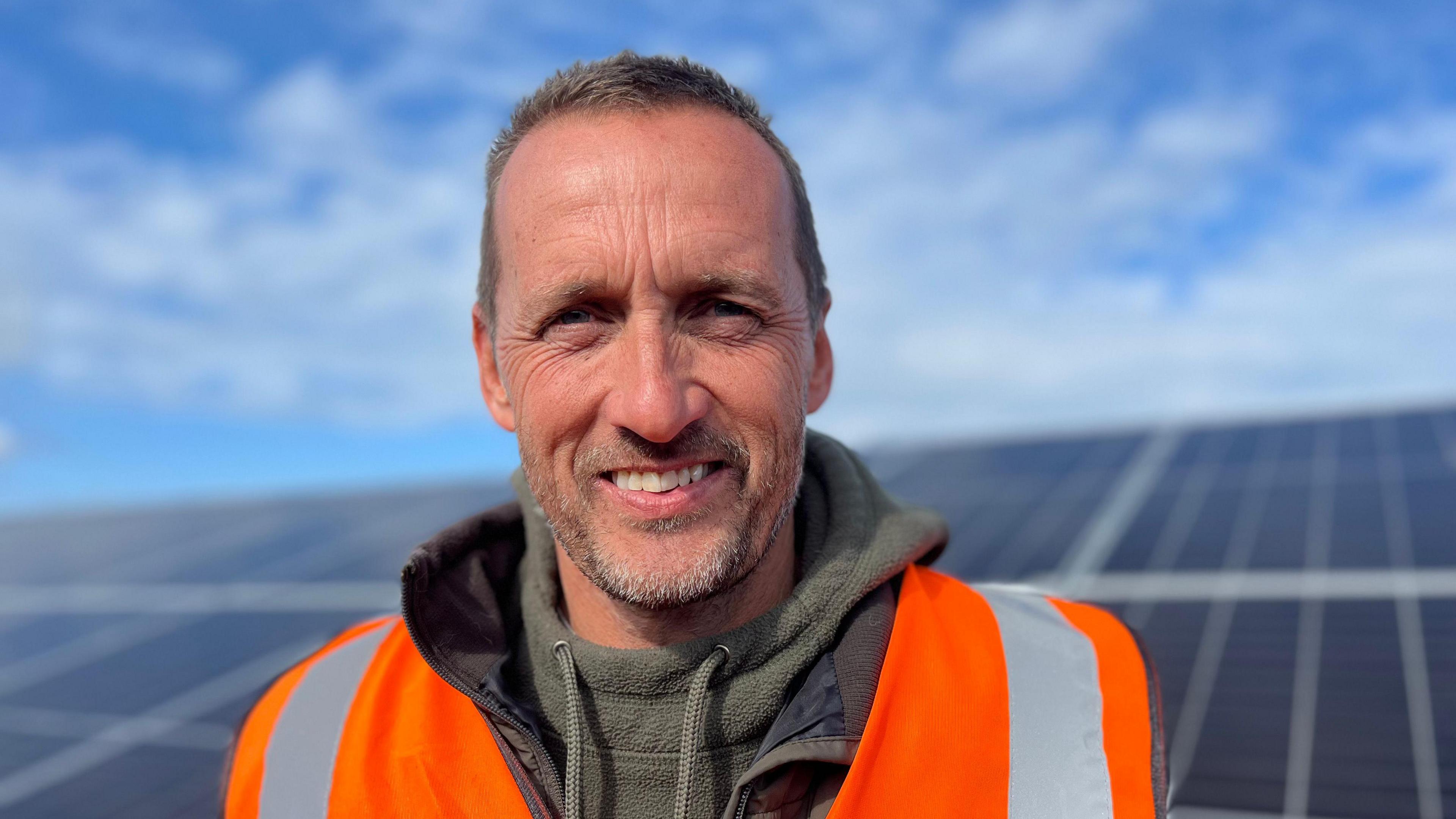 Jeremy Hughes is a white man with grey hair smiling into the camera. He is wearing a green hoodie underneath an orange high-viz vest. The background is an out of focus solar panel and blue sky with clouds.