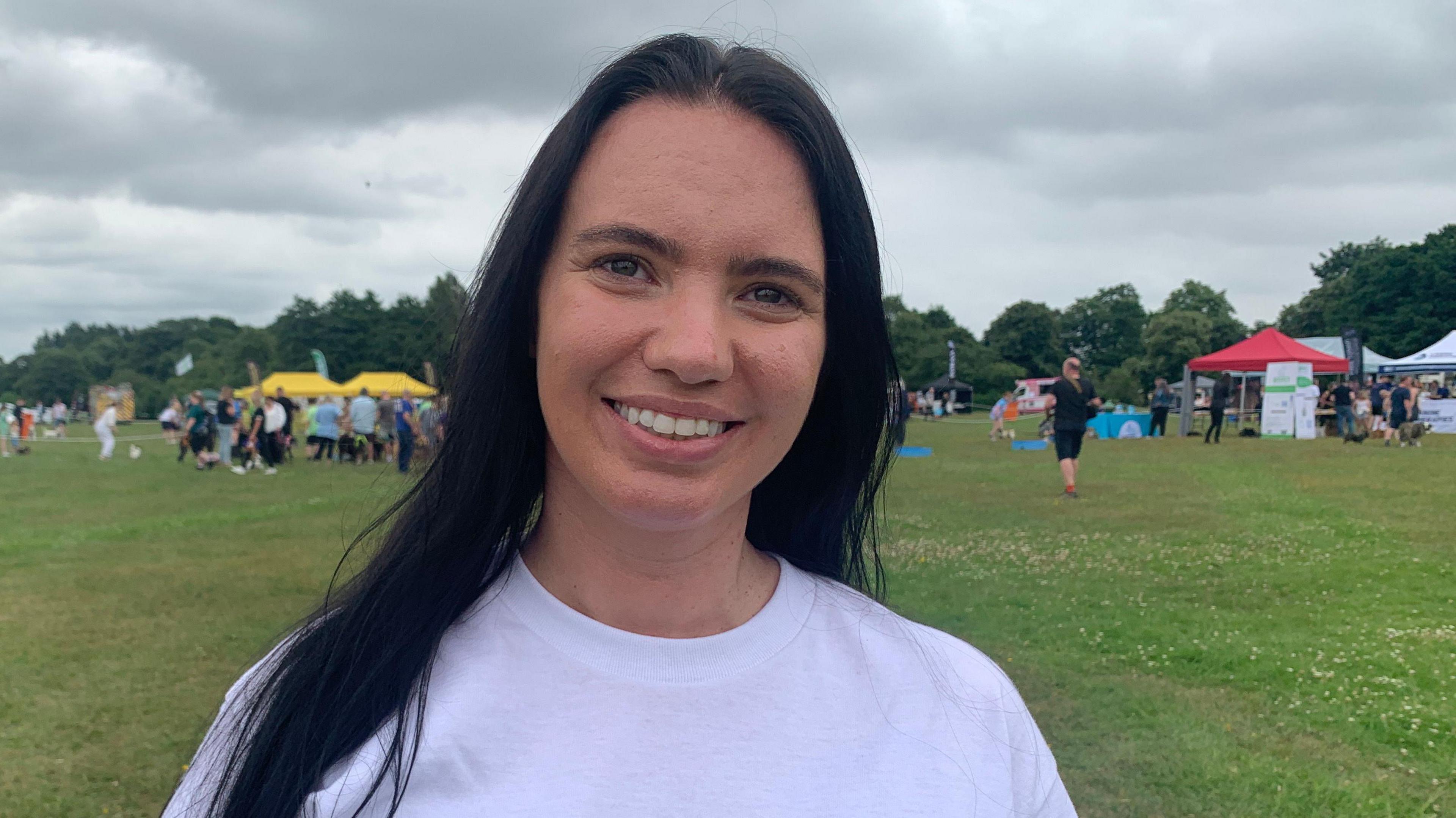 Hannah Carter with long dark brown hair wearing a white t-shirt standing in a field during a festival for animal charities in a park. She is smiling.