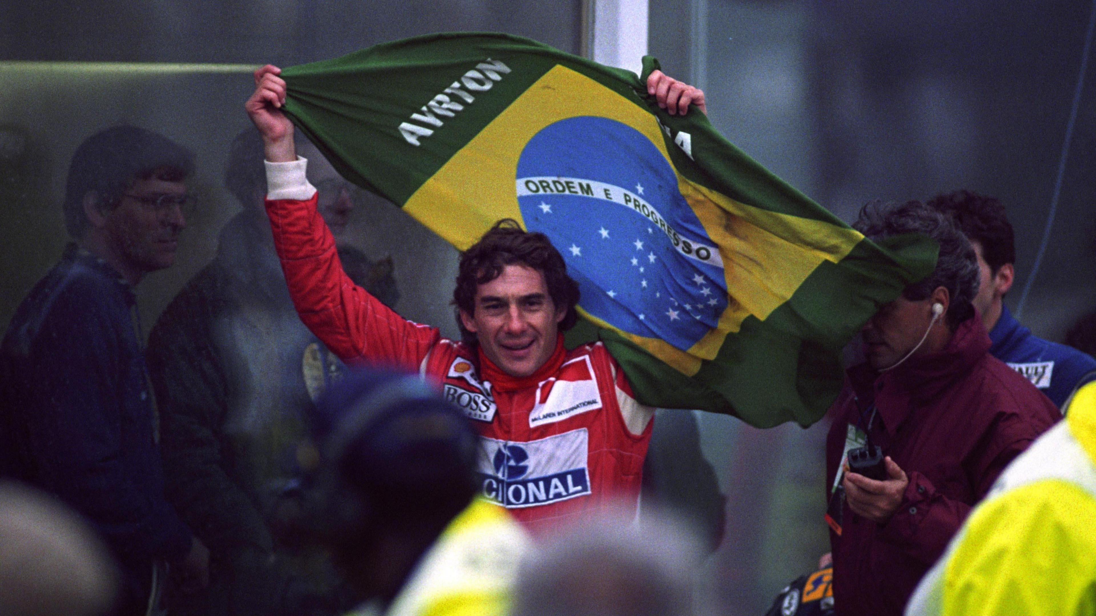 A man in a red racing suit holds up a Brazilian flag