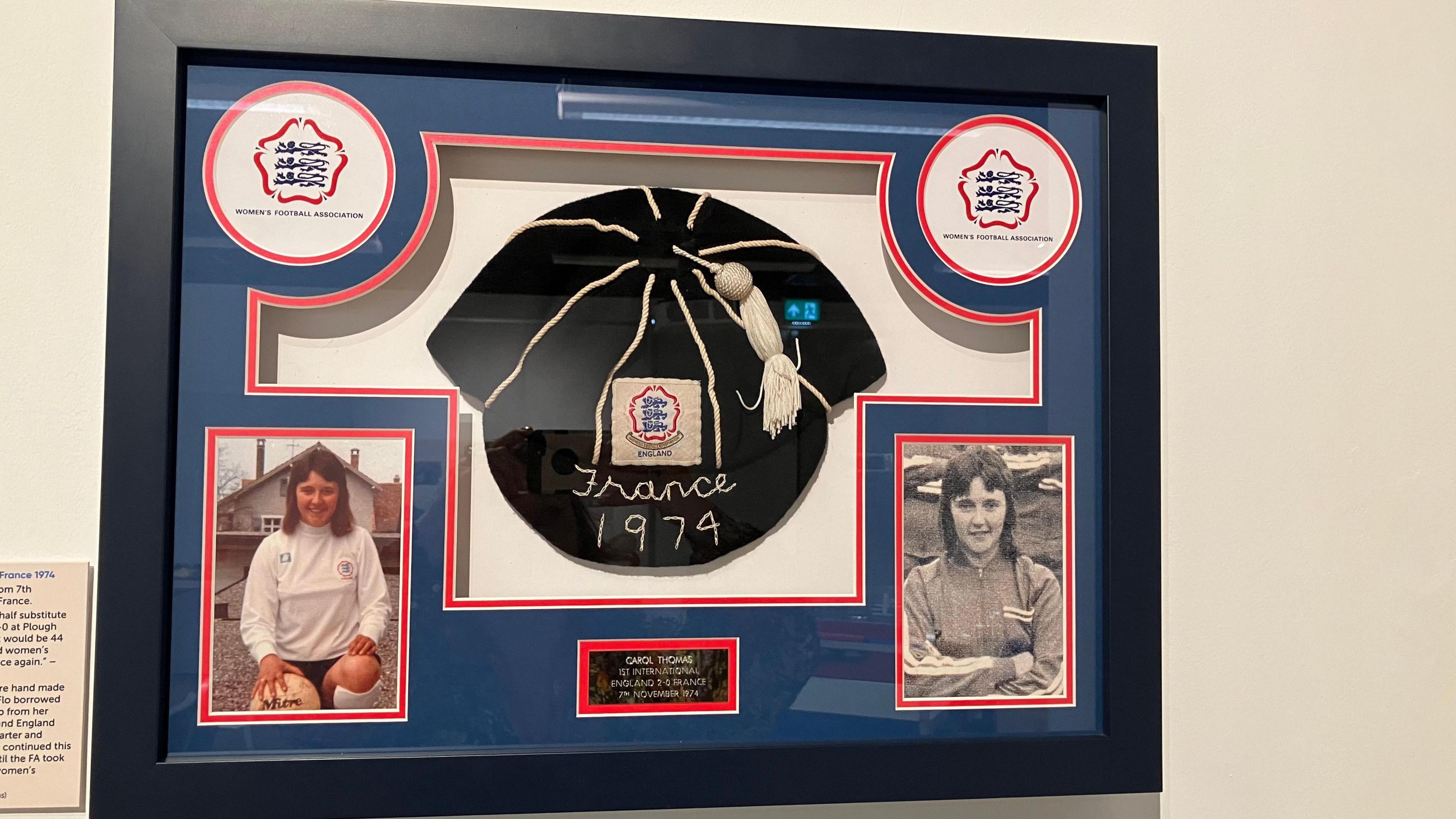 An England 'cap' created by Flo Bilton for Carol Tomas on display - it is black with a white tassel and the Three Lions logo, it reads "France 1974" on the front. The display also features photos of Carol when she was a young player. 