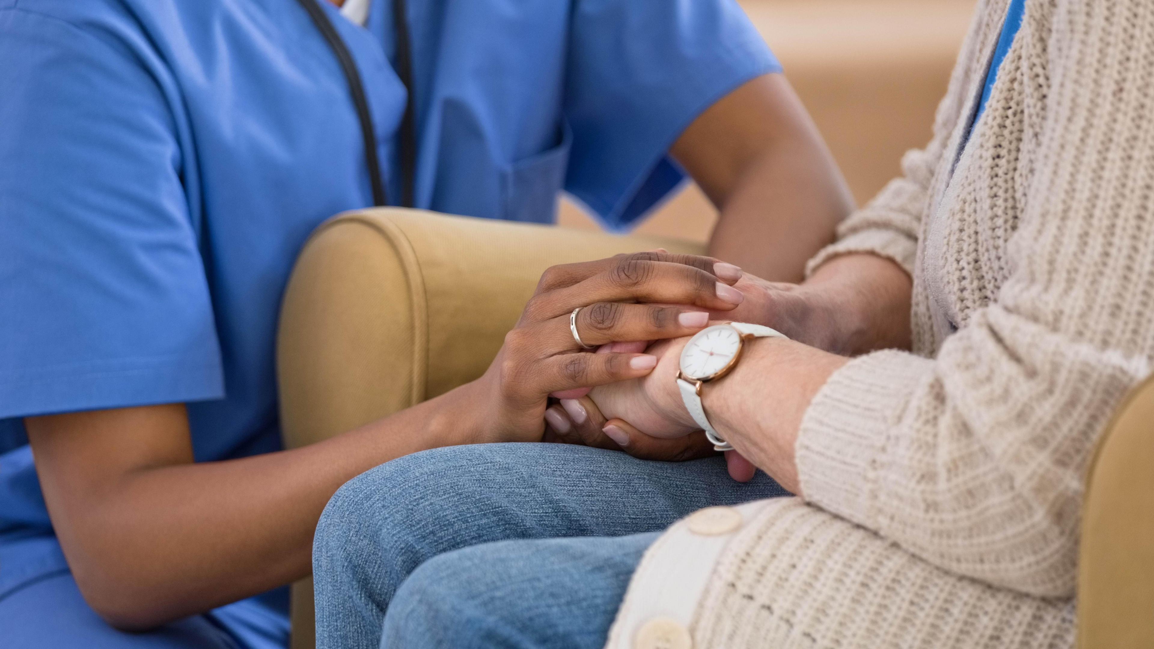 This close up image show a woman in a blue uniform crouching to hold hand of a woman sitting in a chair. The woman wears a long beige cardigan and blue jeans and she has a cream watch on her left arm. 