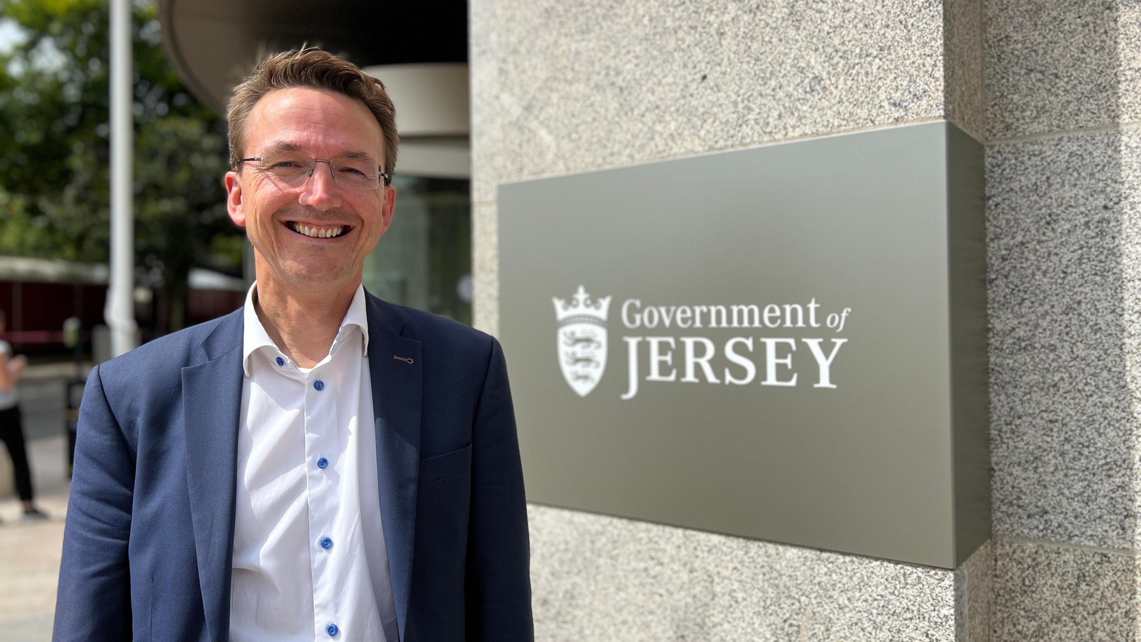 Kirsten Morel stand in front of a building with a sign saying "Government of Jersey". He has short brown hair and is wearing glasses, a white shirt and a blue blazer.