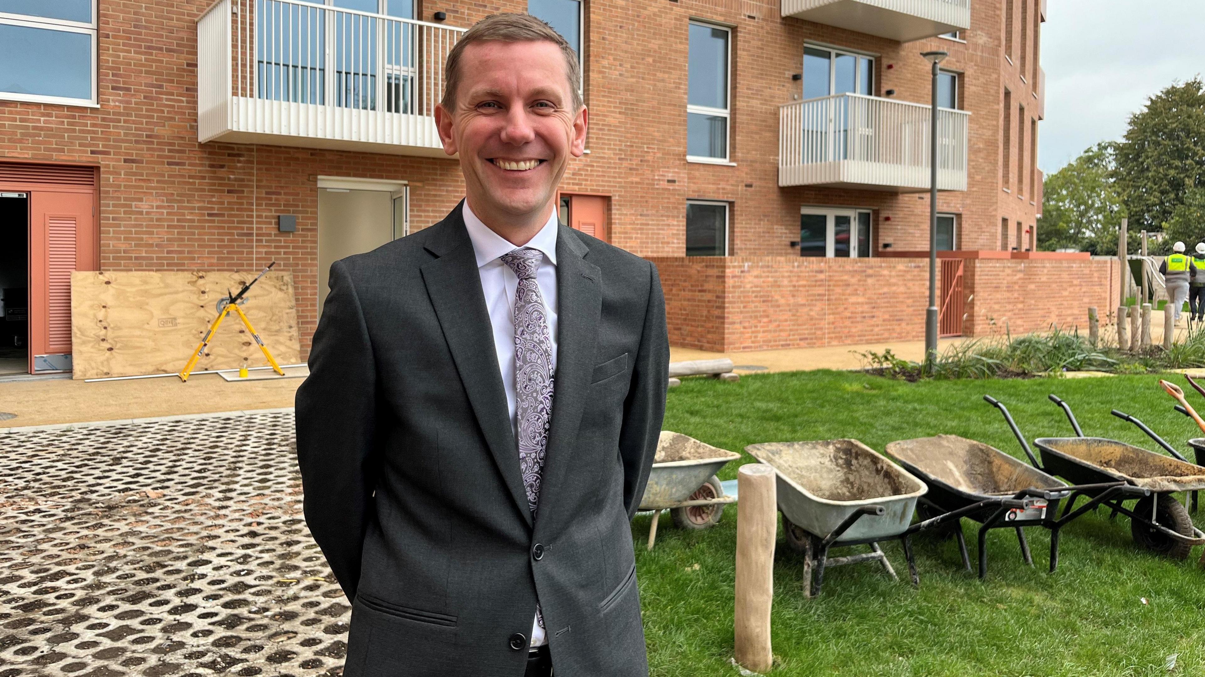 Tom Bruce is seen standing in front of at least four wheelbarrows on the site of the New Road Triangle development in Feltham. He is wearing a suit and pictured with a big smile.