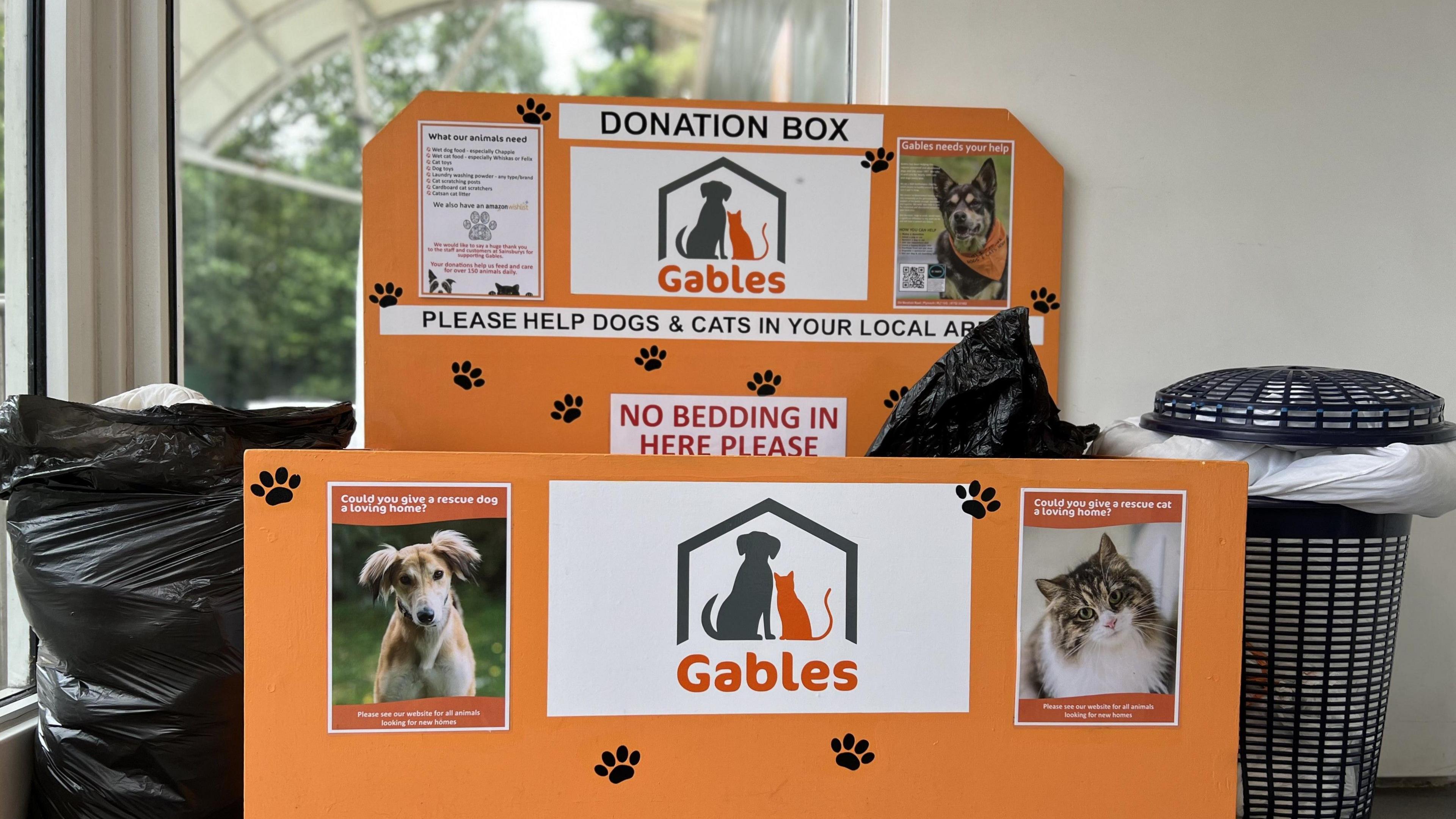 The Gables donation bin. It is orange and has pictures of dogs and cats. At the centre is the Gables logo with the word 'GABLES' below it. There is a black bag, with a duvet inside beside the box, and a basket with other bedding in it on the other side.