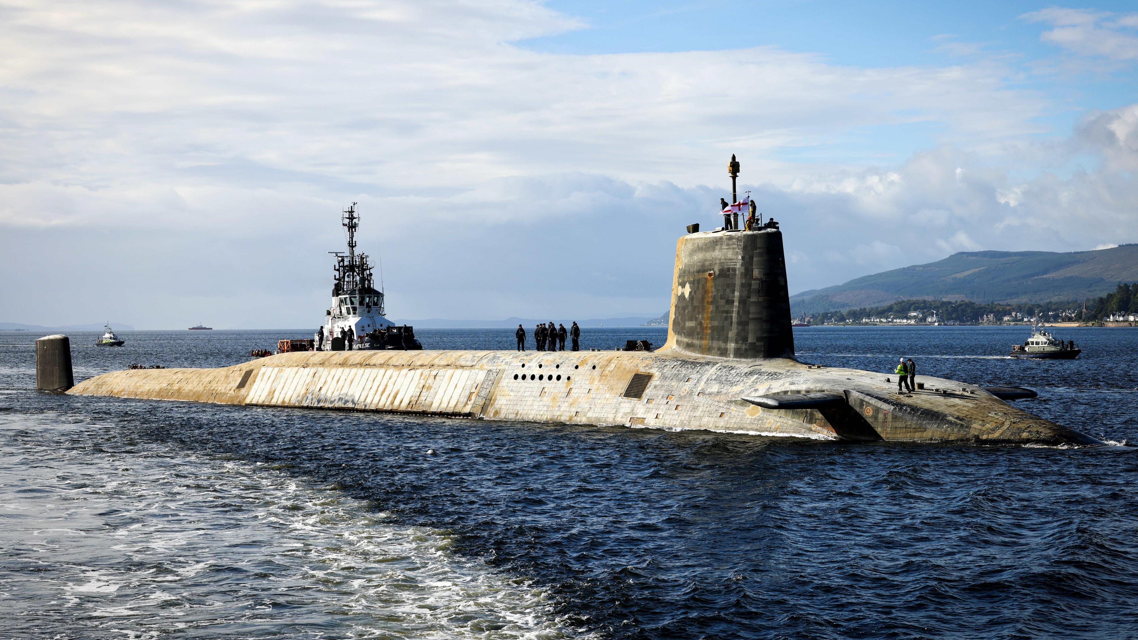 HMS Vanguard, a large grey submarine, in the Clyde with sailors standing on top. A tug boat can be seen alongside.