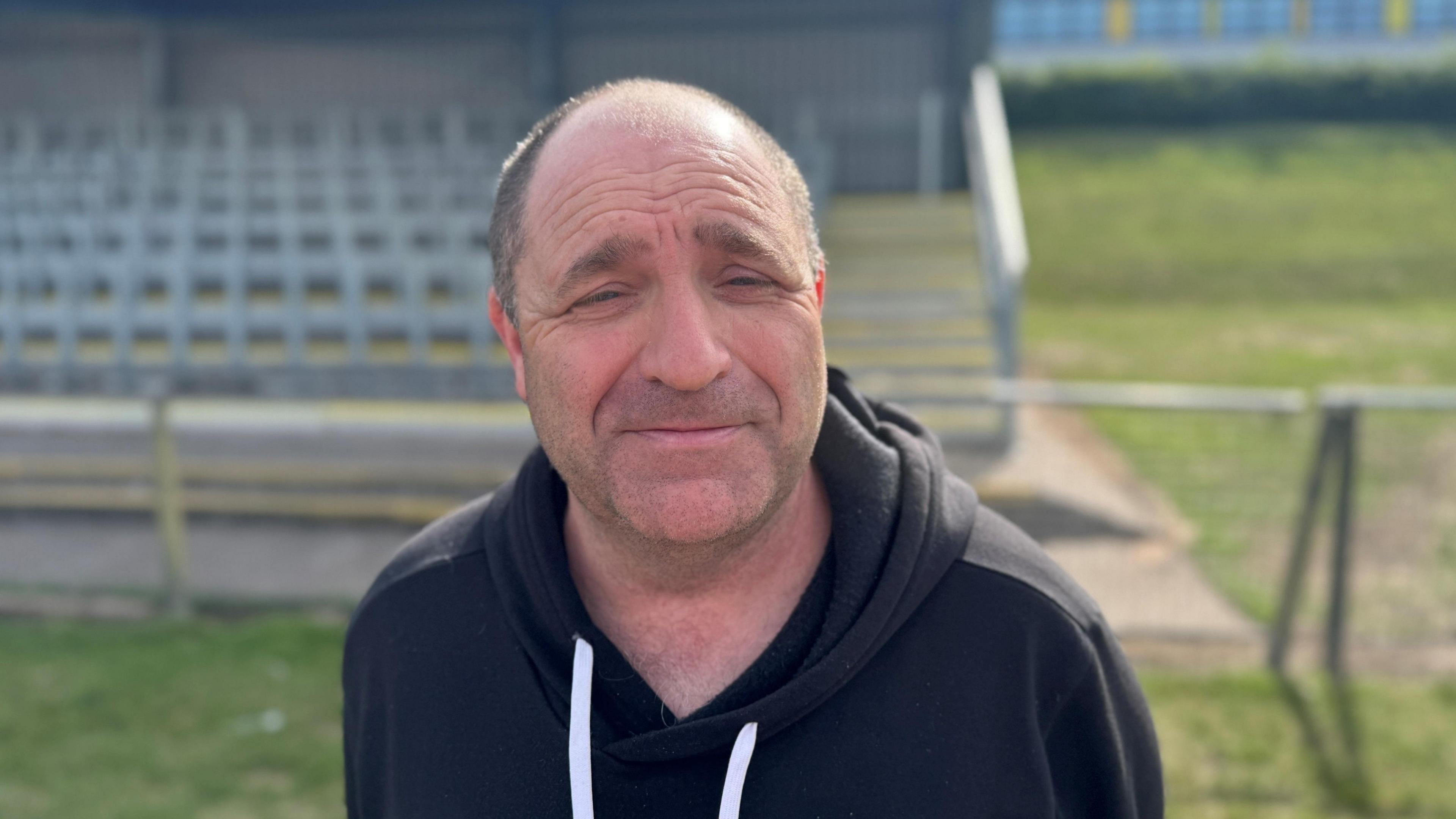A man with short dark hair and a dark tracksuit on a grassy area