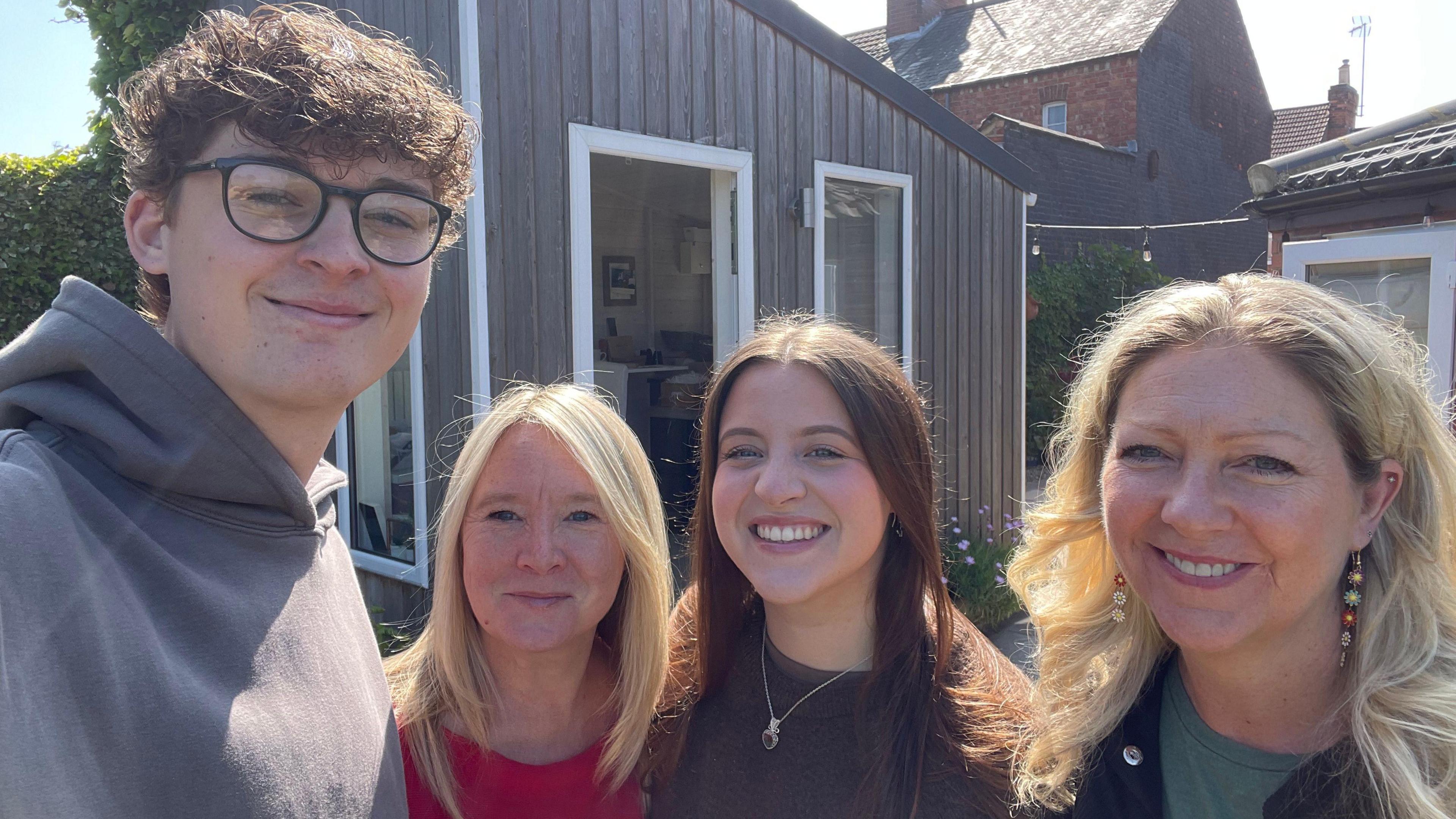Hugh, Cara, Leah and Lesley, all standing outside, in a garden, by a small building, all looking at the camera and smiling. 