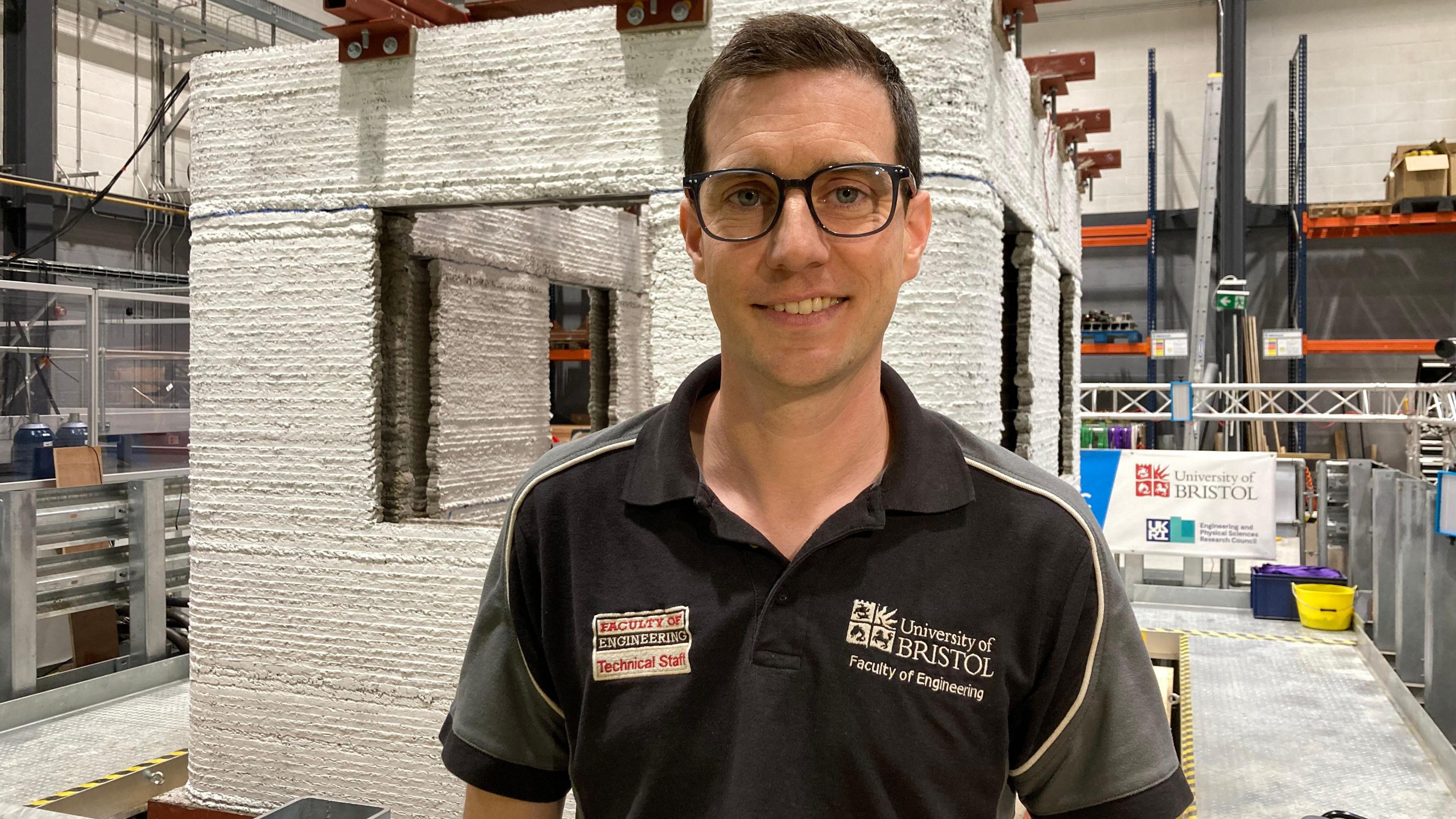 A white man with brown hair and glasses, wearing a University of Bristol 'Faculty of Engineering' polo shirt is smiling at the camera. He is standing in front of a large 3D structure.