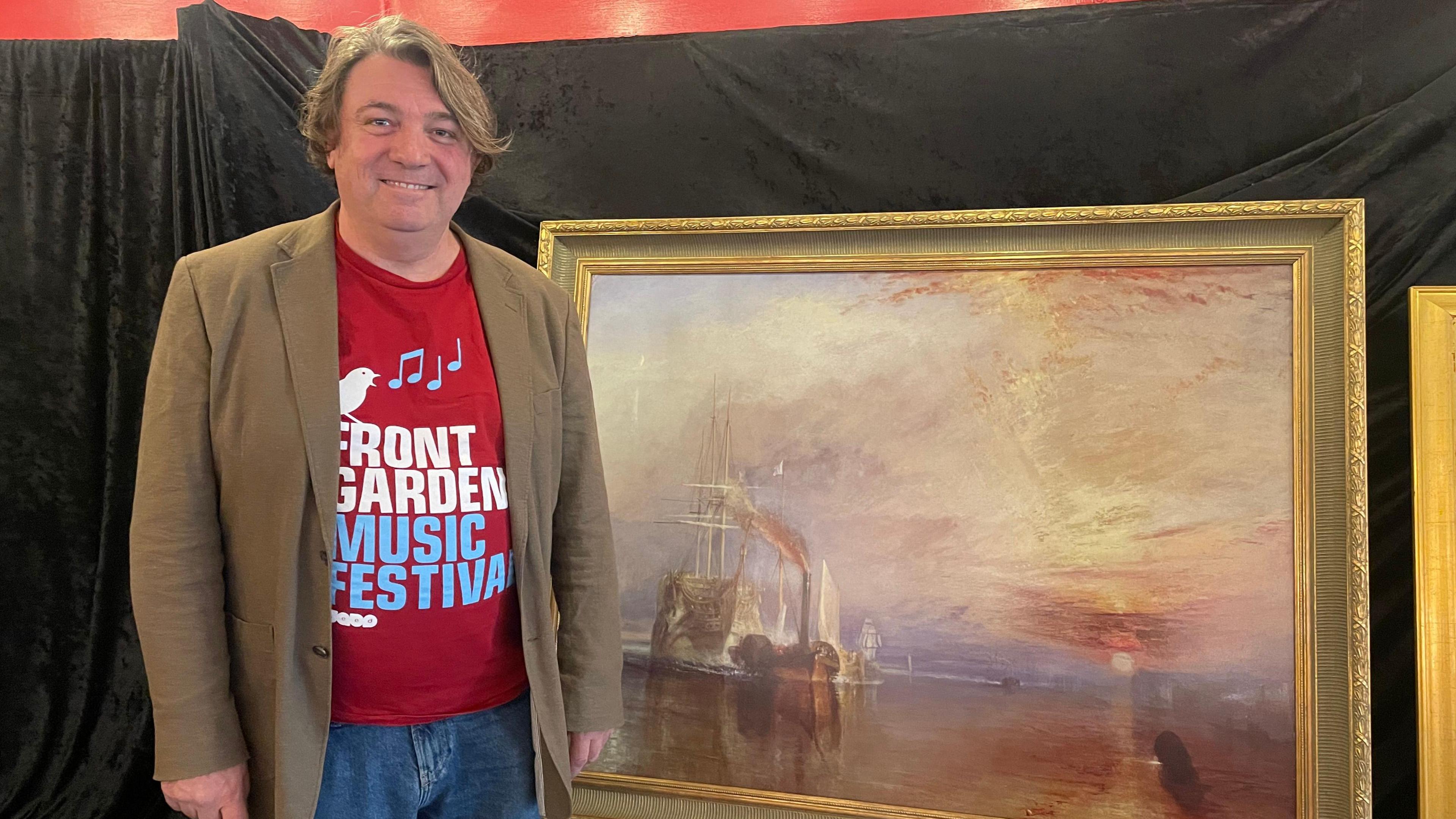 Scott O'Hara stands smiling beside a Turner painting. He is wearing a light brown jacket and a red T-shirt with red and blue writing on it