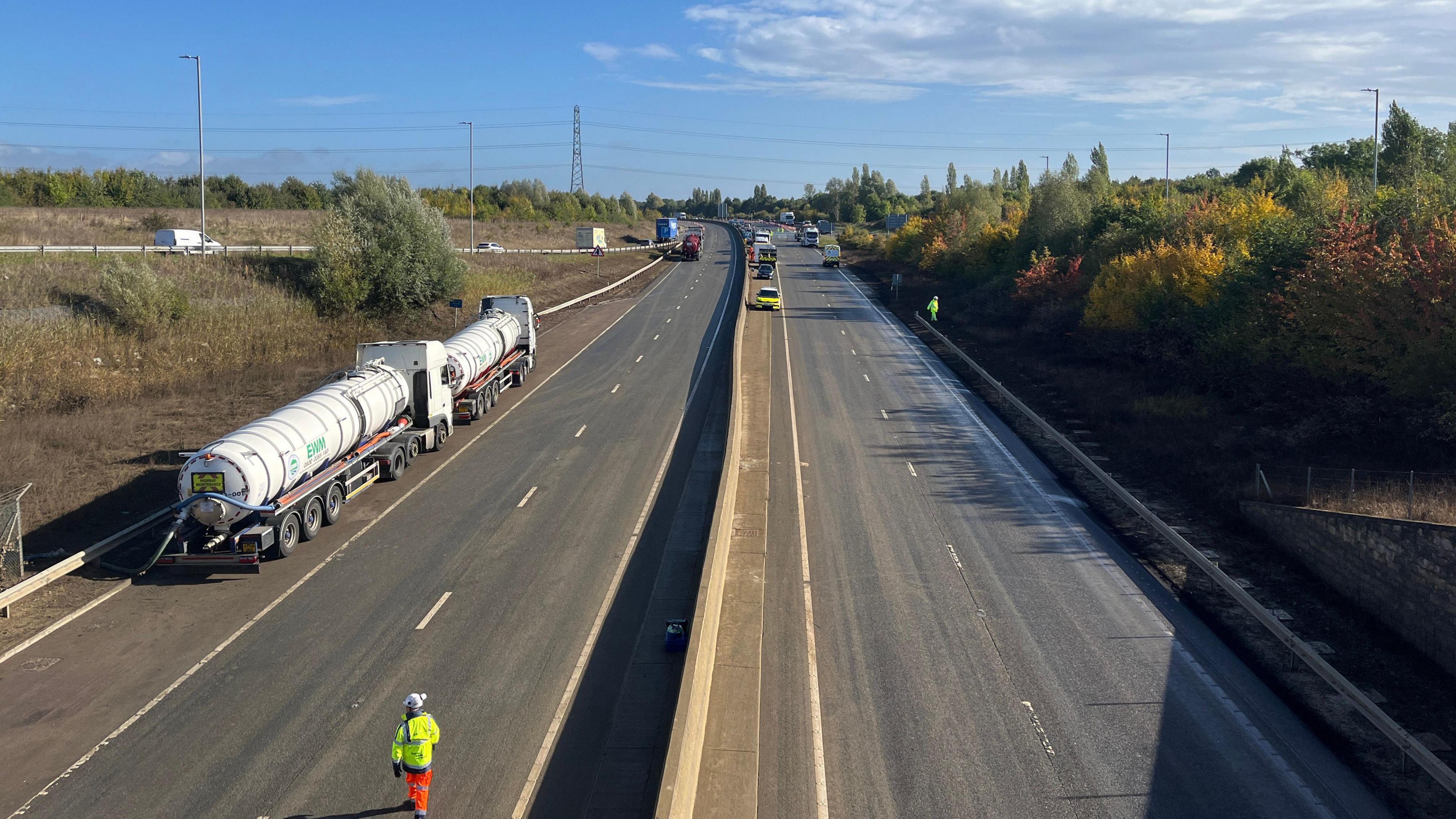 Highways crews are pictured on the A421. Tanked can be seen parked on the hard shoulder at the side of the road with large pipes coming from them. Workers in high-vis jackets can be seen on the road while there are vehicles in the distance heading onto the road via a slip road.