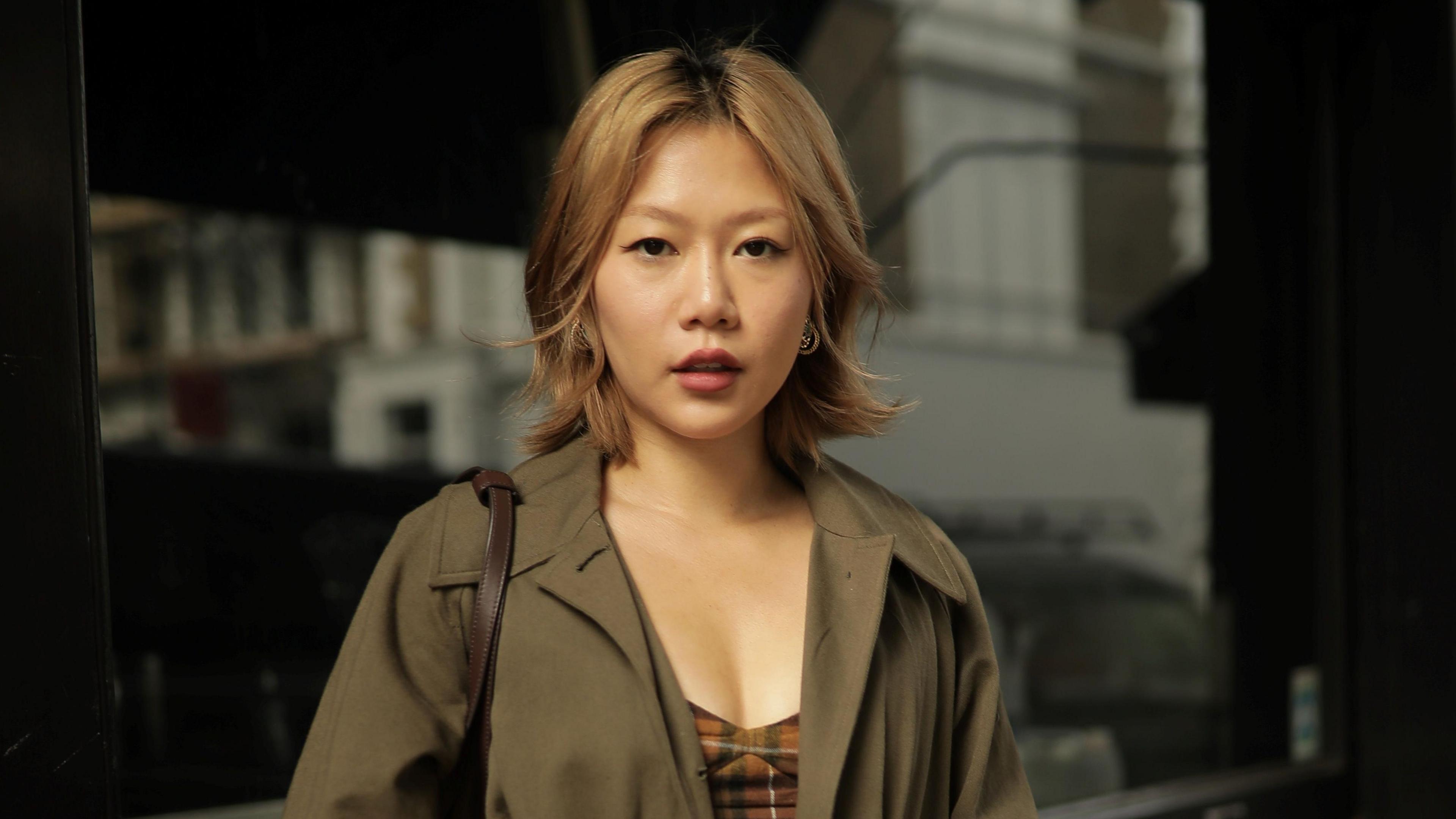 Andrea Cheong pictured outside a shop window. She wears a green overcoat and an orange tartan-patterned top. She has a blonde bob and looks at the camera. 