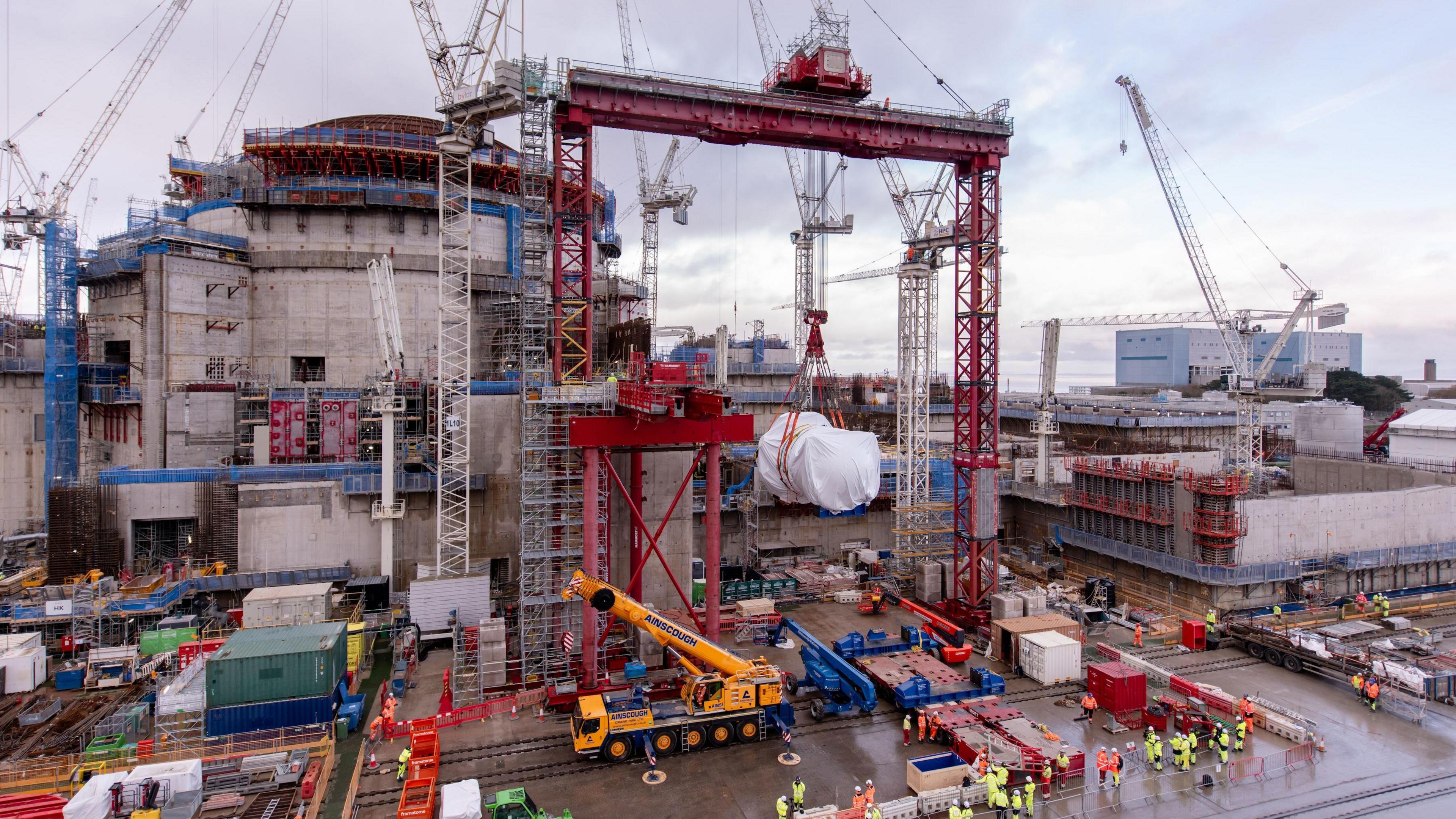 A big cylinder shrouded in white wrapping is lifted up on a big red crane. It stands in front of a large concrete building, with a steel brown domed roof. Dozens of workers in high vis are on the ground, and there are several other cranes all around.