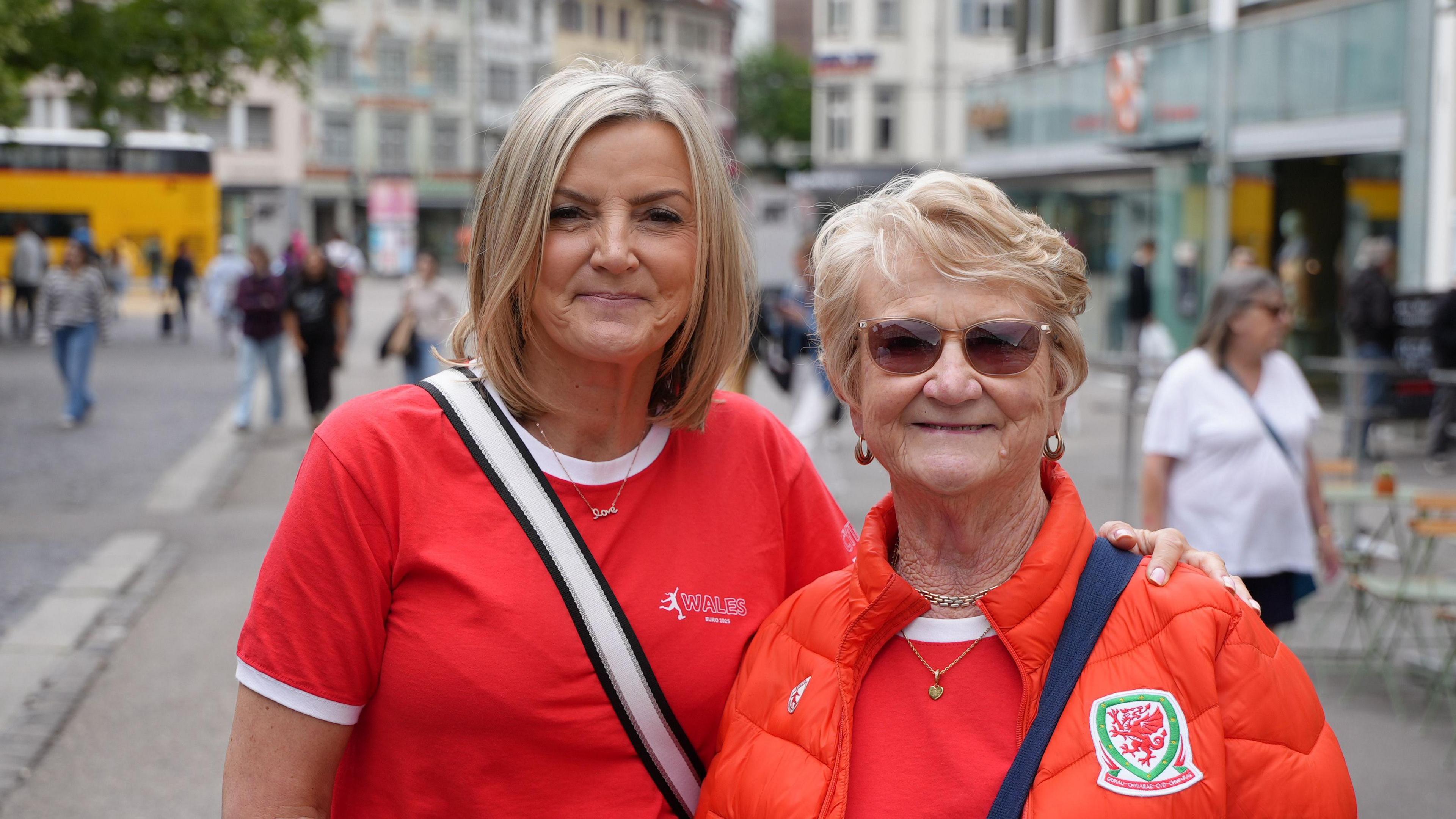 Two women looking at the camera, both have a variation of a red top on. The woman on the left has her blonde hair cut into a bob. The woman on the right is slightly older and has sunglasses on.