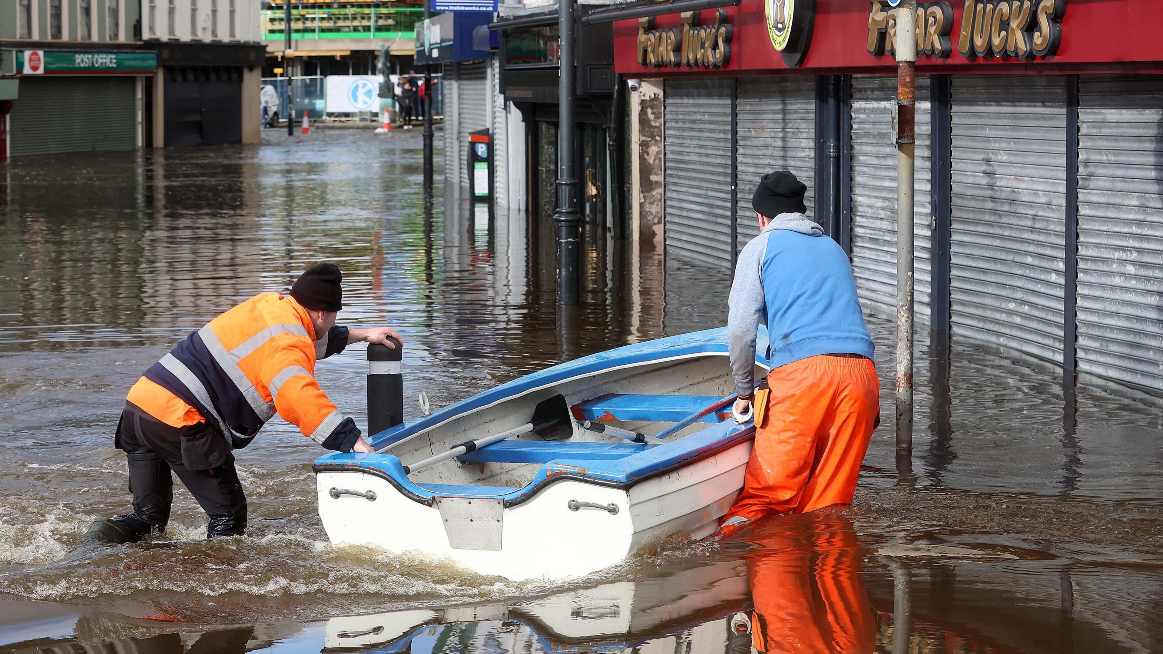 Two men in high-vis gear pushing a small row boat through knee-high water in the middle of a city street with shops on either side.