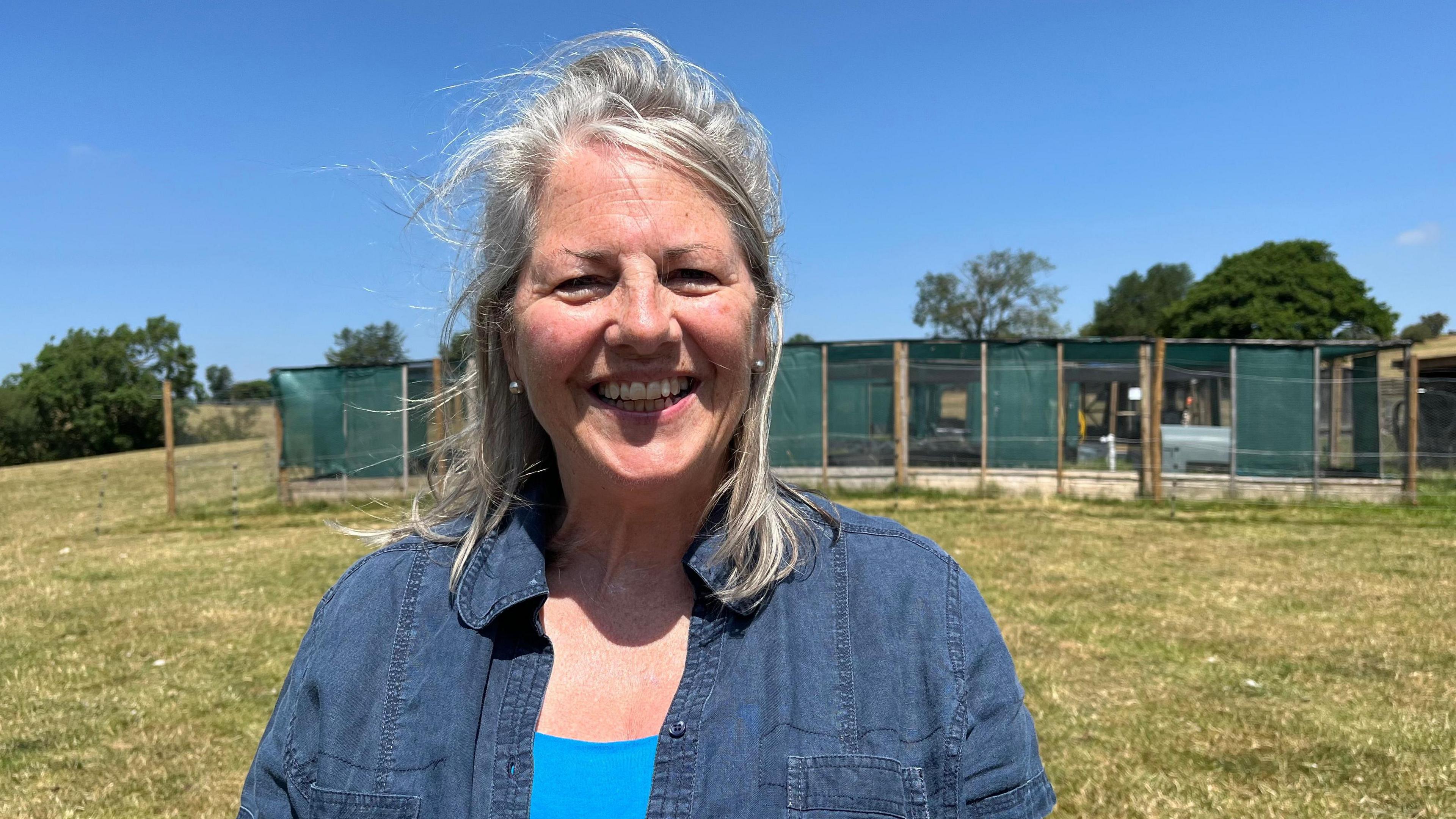 Perkins photographed looking into the camera and smiling. She is wearing a dark blue denim shirt with a bright blue vest underneath. She is stood on a large grassy field, with pens for the curlews behind her built out of chicken wire, wooden posts, and green material. The sky is clear and blue and there is strong sunshine. 