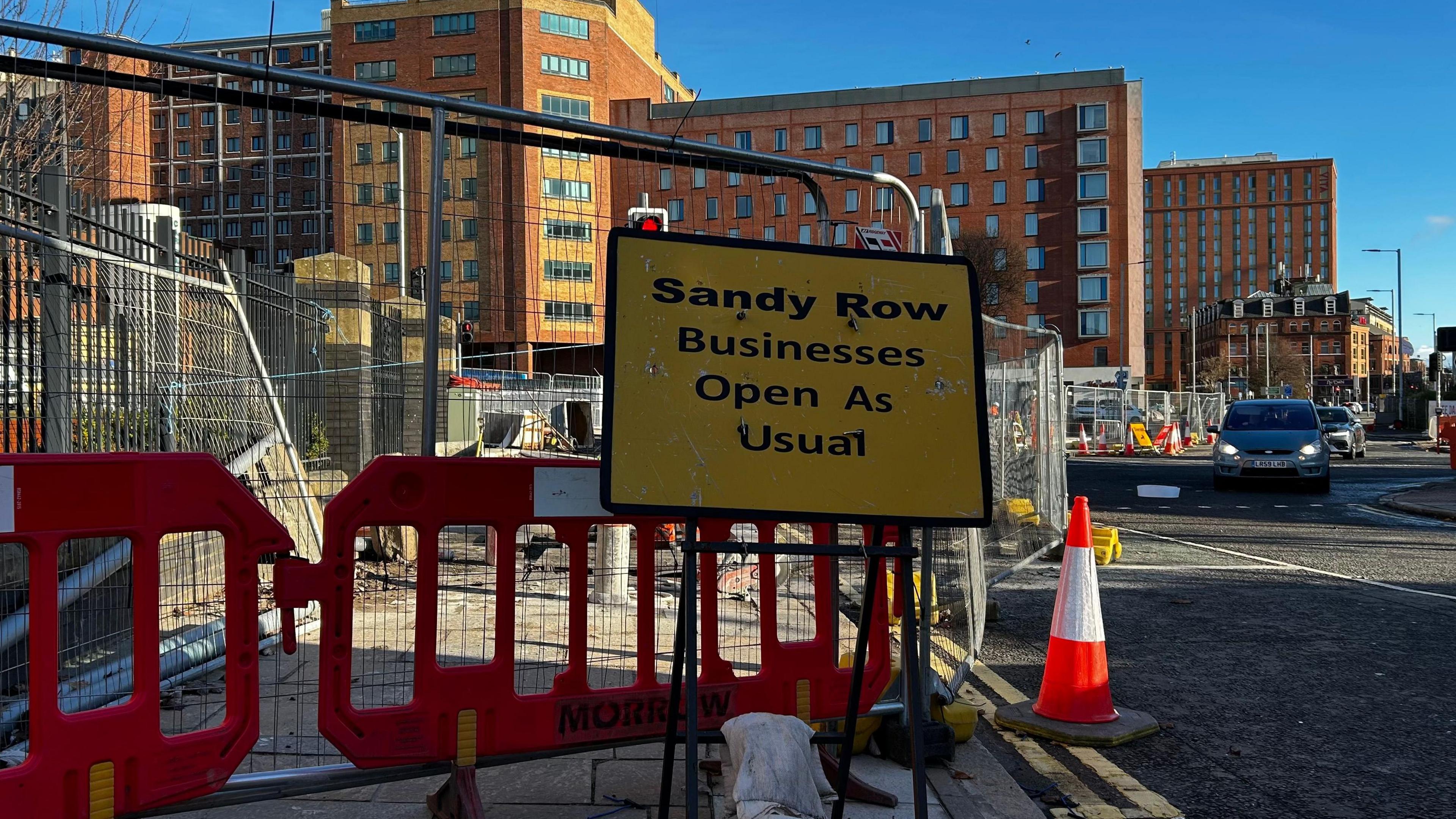 A picture of a yellow road work sign in front of a fenced off area. It says 'Sandy Row Business Open As Usual'. A traffic cone can be seen beside the barriers. Red brick buildings can be seen in the background as can cars on the road which is open to the side of the frame.