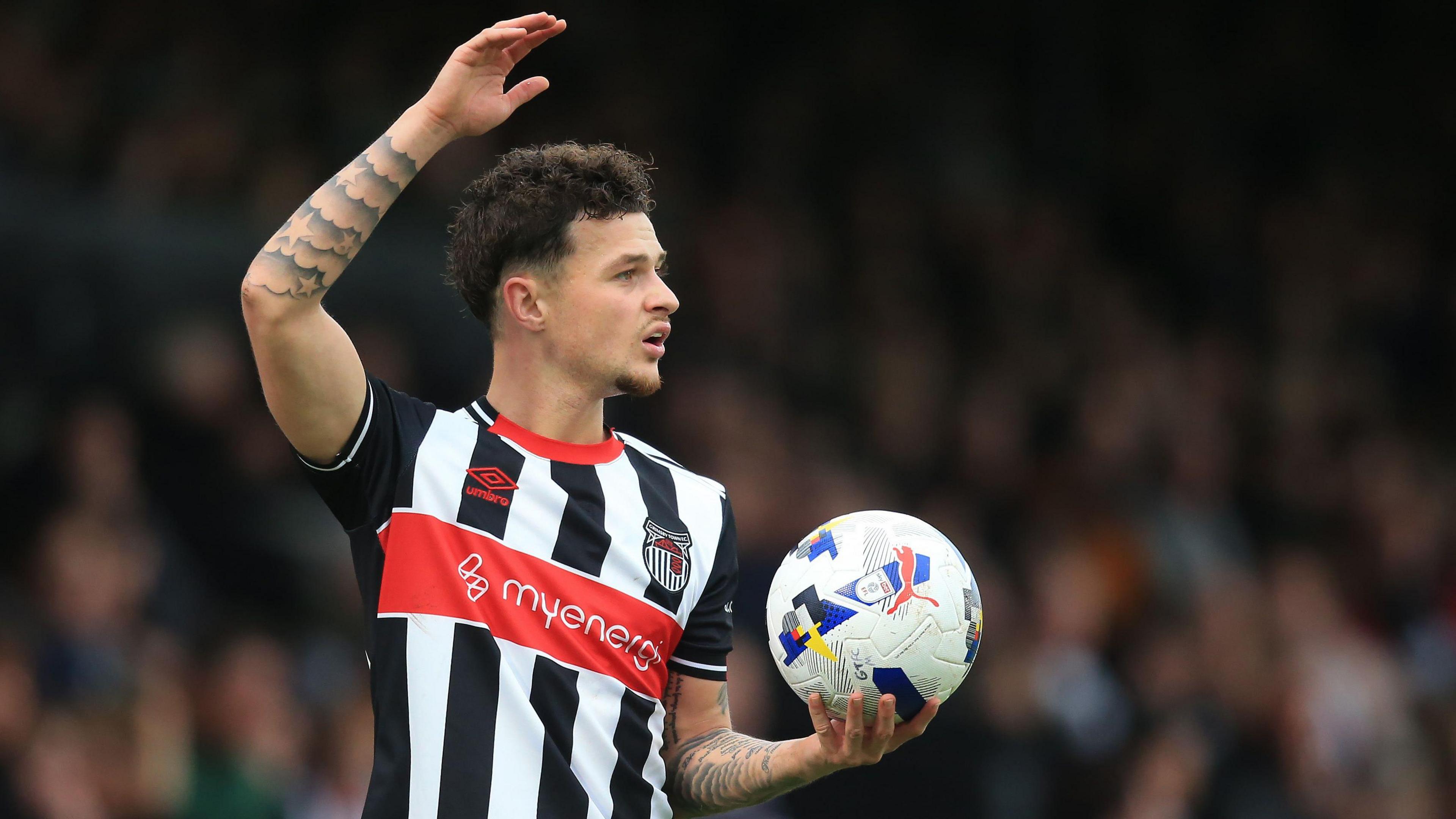 George McEachran, in black and white Grimsby kit, holds up his right arm with the ball held in his left hand.