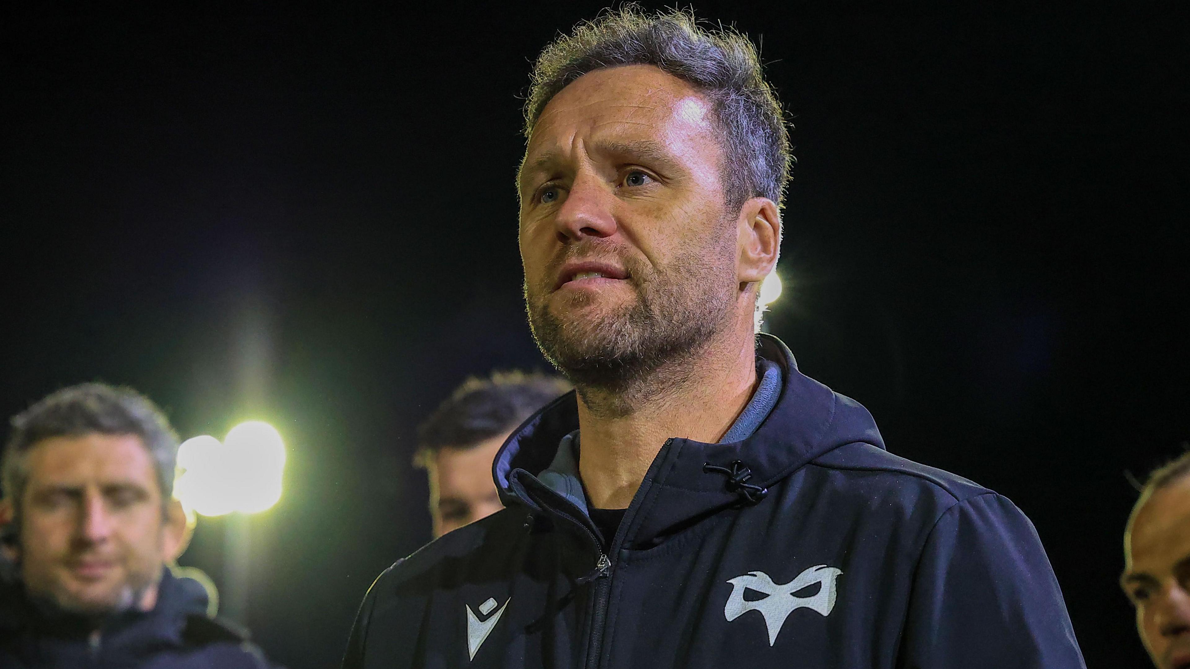 Ospreys head coach Mark Jones stands in front of other Ospreys' staff in a floodlit ground