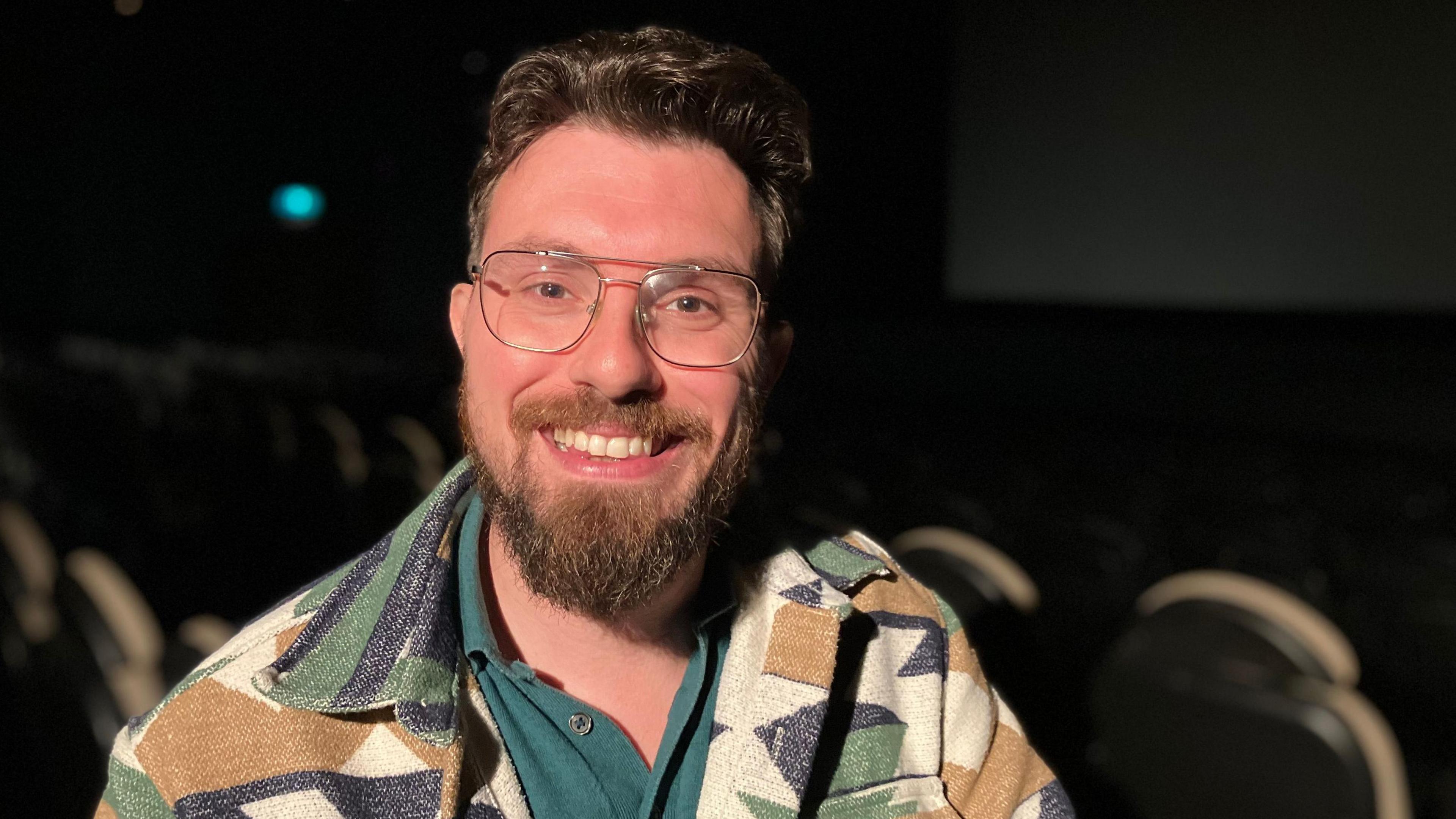 Hendrik Harms is interviewed in a cinema. There are seats behind him in the theatre with a screen in the background. He is wearing a green top and a jacket and wearing glasses. He has dark brown hair and a beard.