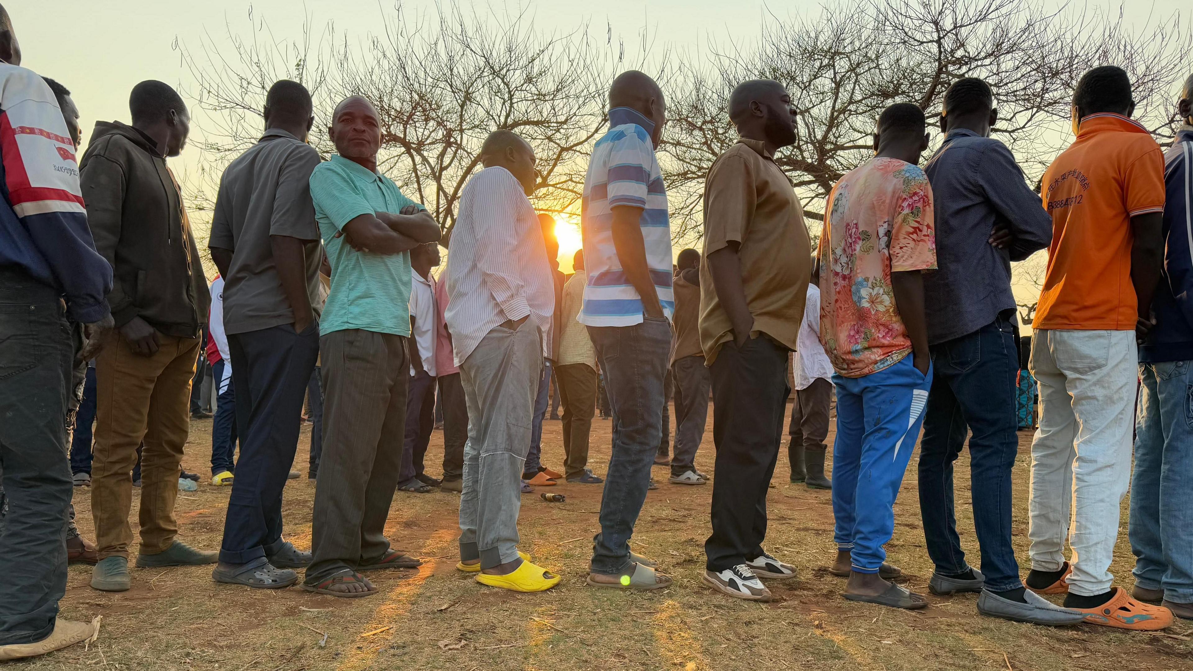 A wide shot taken from the side of a queue of people waiting to vote on a dusty, dry field. The rising sun is just poking through the bodies in the middle of the queue. 