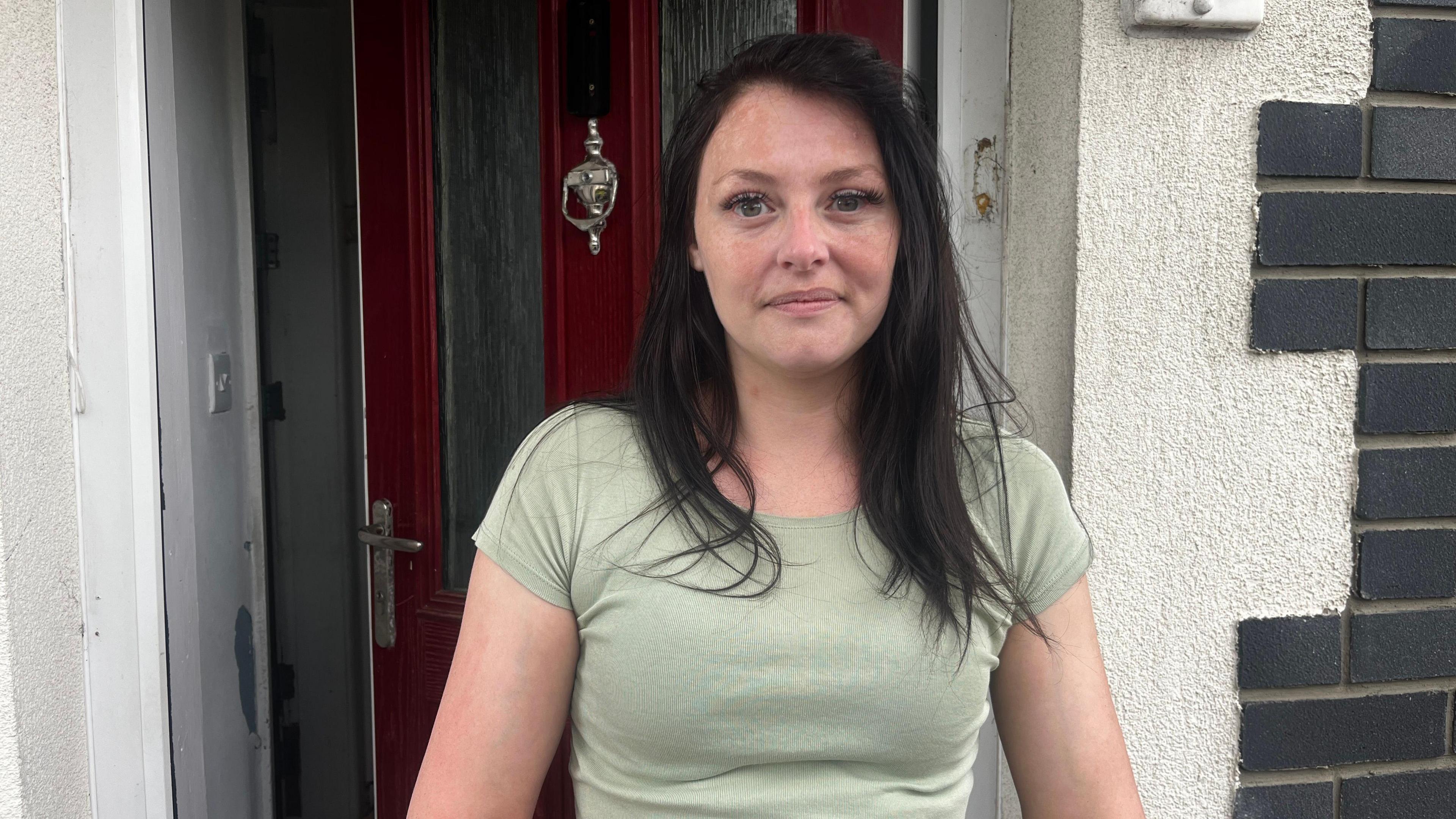 Melissa Taylor, 31, who has long straight black hair and wears a light green top, looks into the camera in front of a red front door