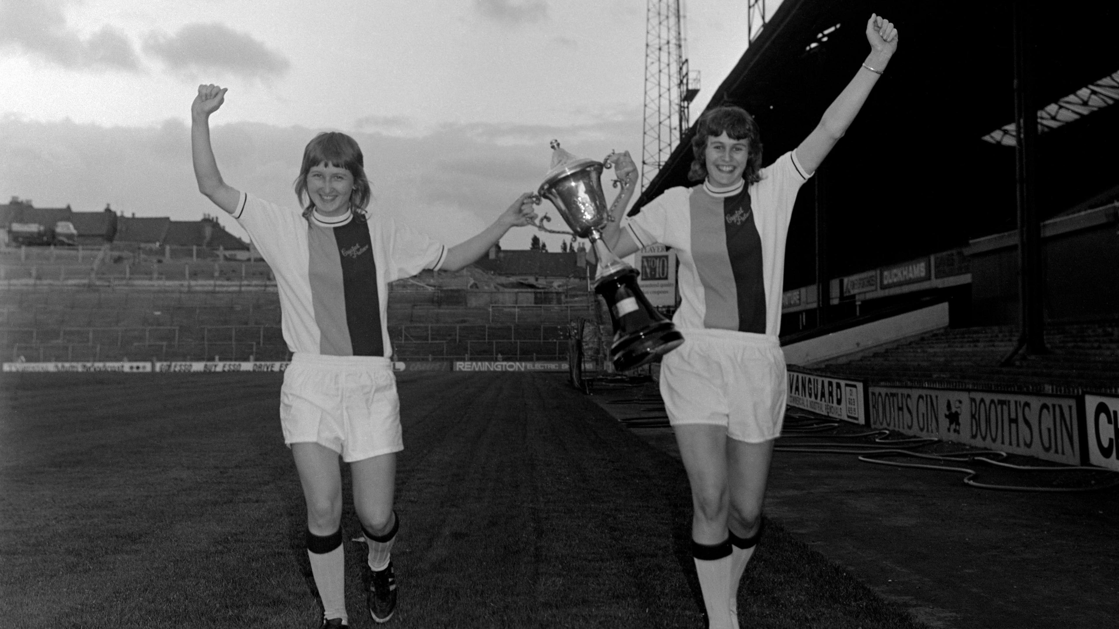 Two Crystal Palace ladies players walking side-by-side on pitch holding a trophy together one hand each and their fists raised.