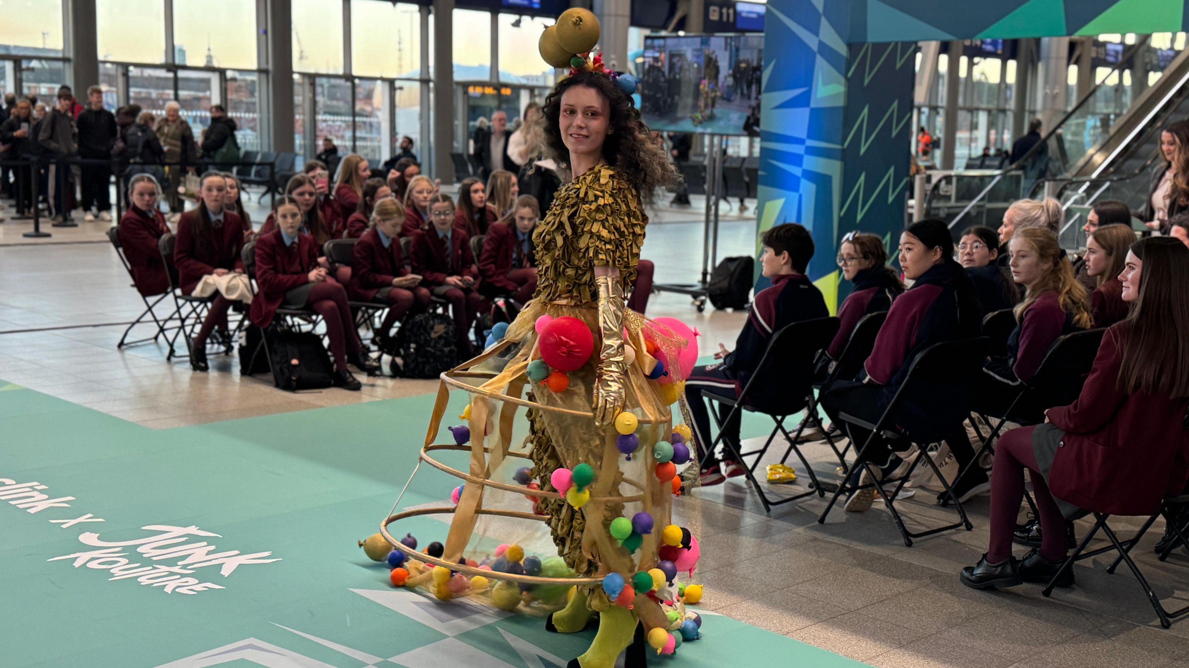 Young person wearing a yellow outfit with old balloons of various colours. The student is walking the catwalk. 