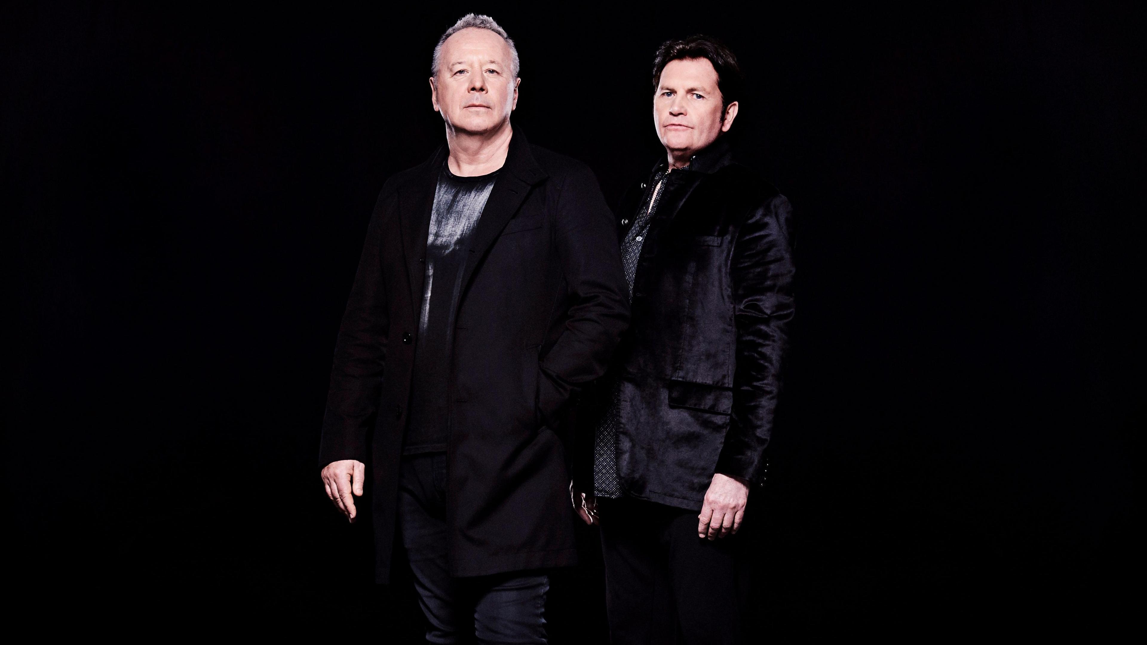 Two men, standing with a black background, looking at the camera. Jim Kerr, is wearing black, with short grey hair, and Charlie Burchill, is also wearing all black. 