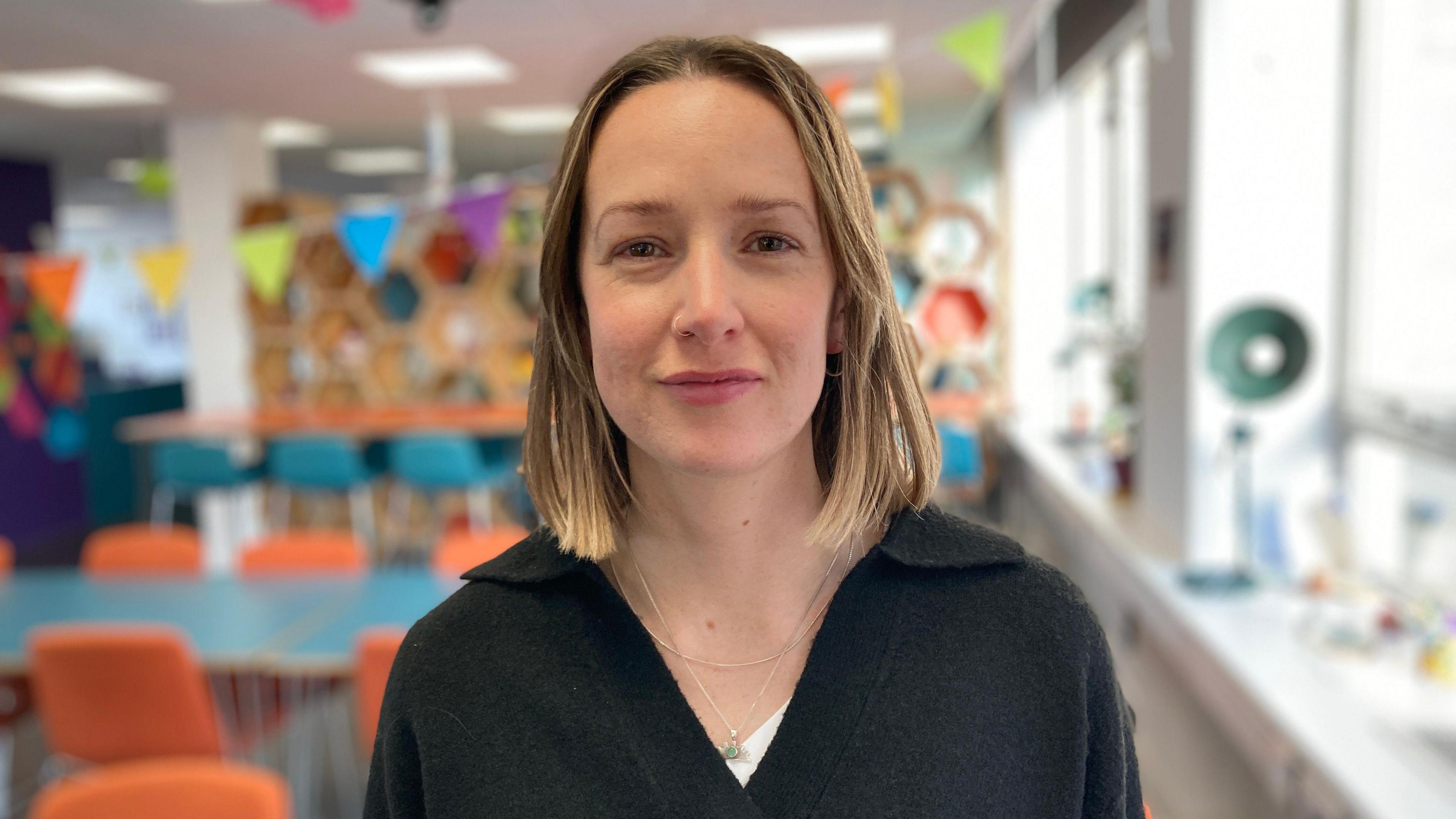 Holly Rooke wearing a black collared cardigan and a thin silver necklace with a small green pendant. She has dark blond shoulder length hair and is smiling at the camera. She is standing in a very brightly decorated classroom with blue tables and orange chair. 