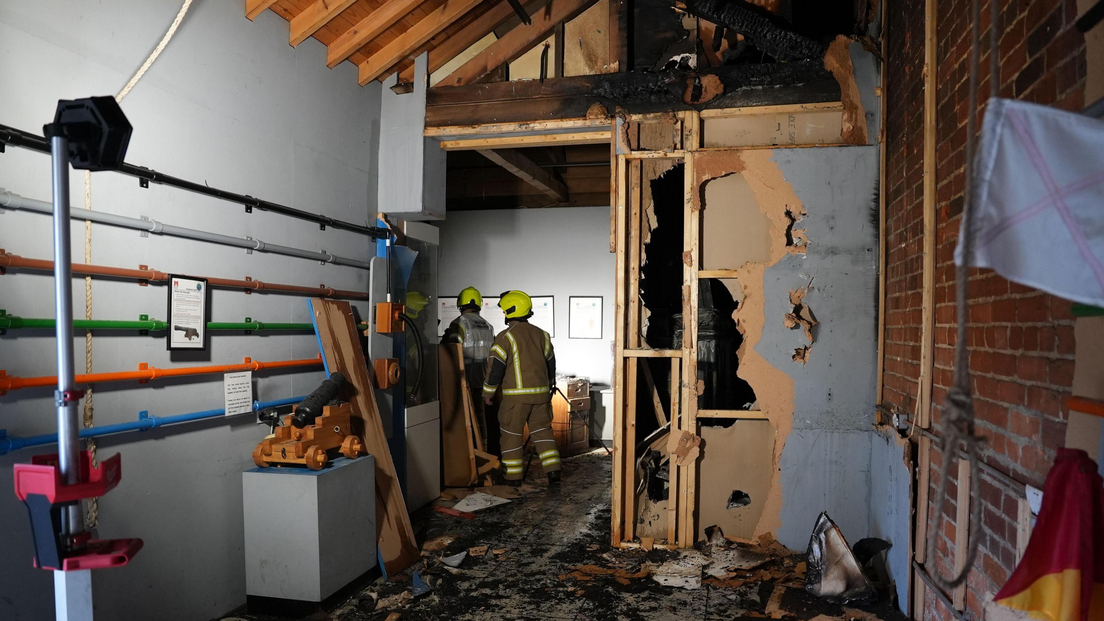 Two firefighters are walking through the museum. To their left are displays but on their right a wall has been damaged by fire. There is debris on the floor and large damaged pieces of wood scattered behind them.