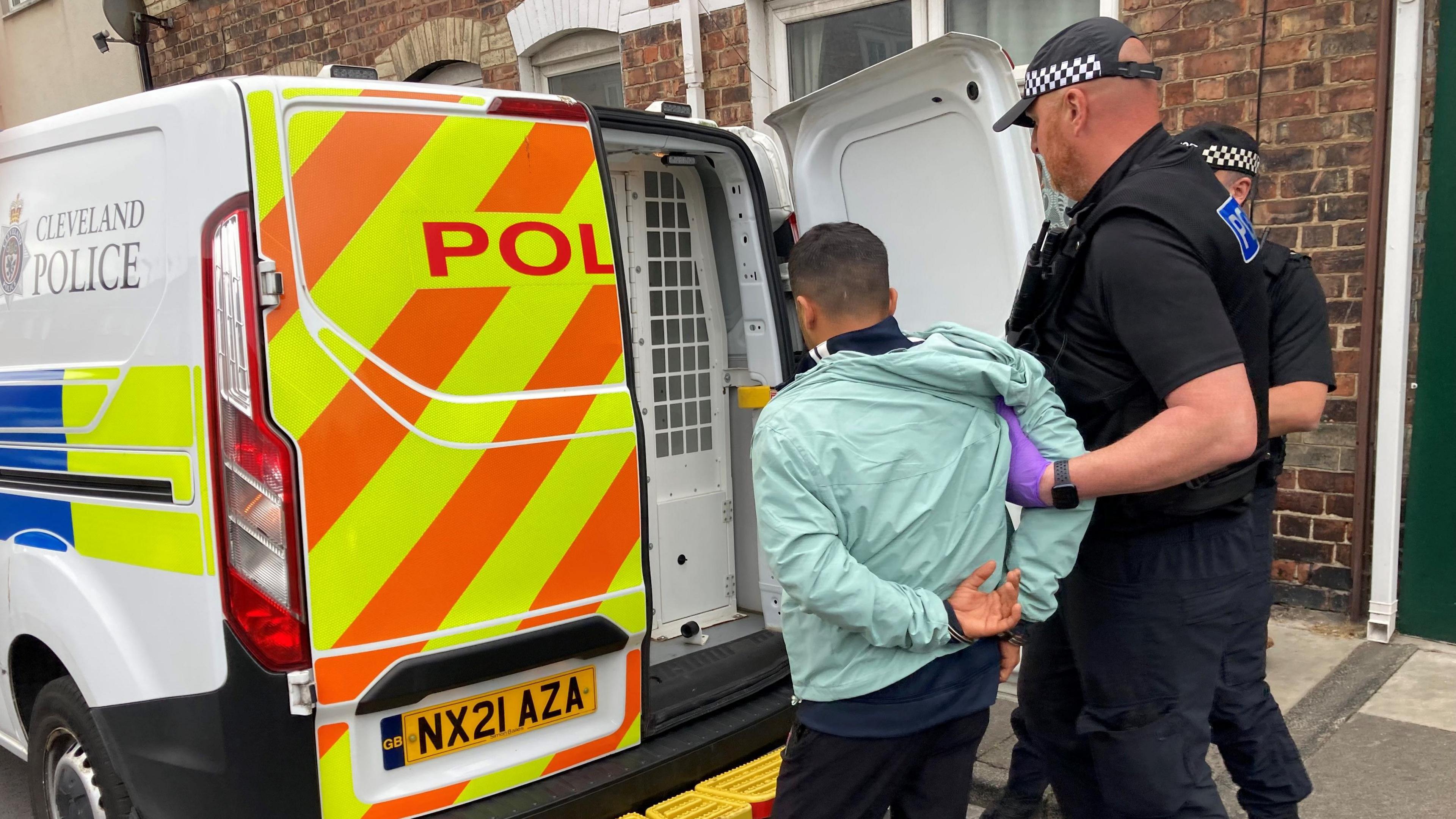 A police van parked outside terraced houses. Two men dressed in all black clothing, with caps on that have police checked patterning on, grab the arm of one smaller man, wearing a light blue tracksuit top and black pants, into the back of the police van.