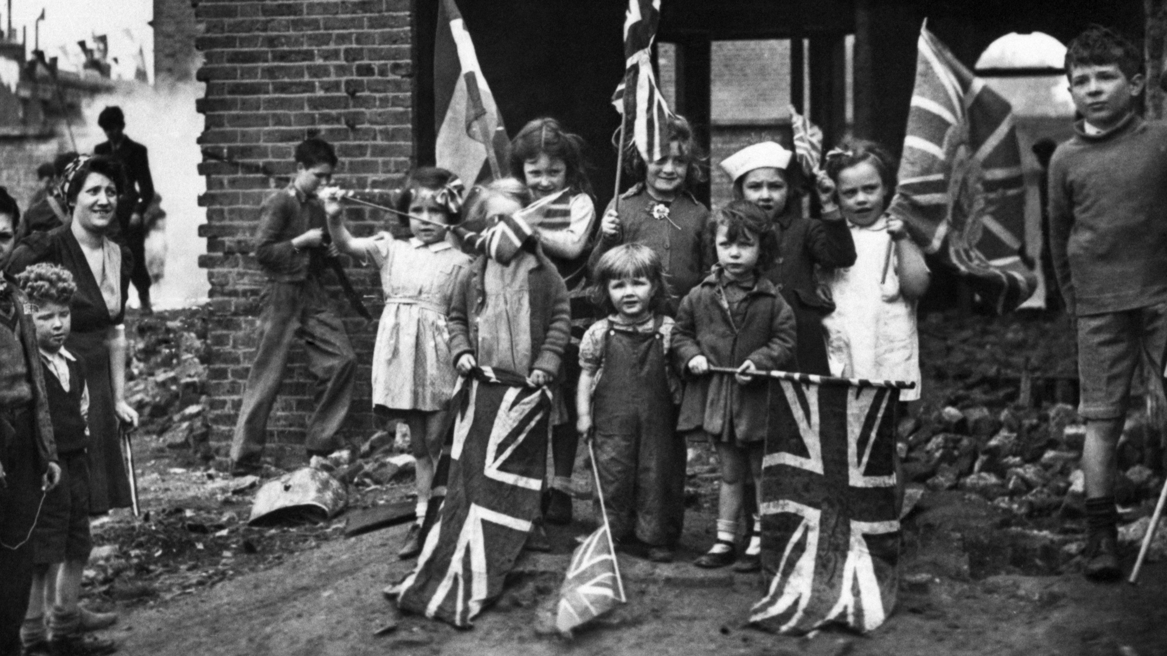 A black and white photo of a group of children holding union jack flags, stood in front of a bombed out building in London. Taken in 1945. 