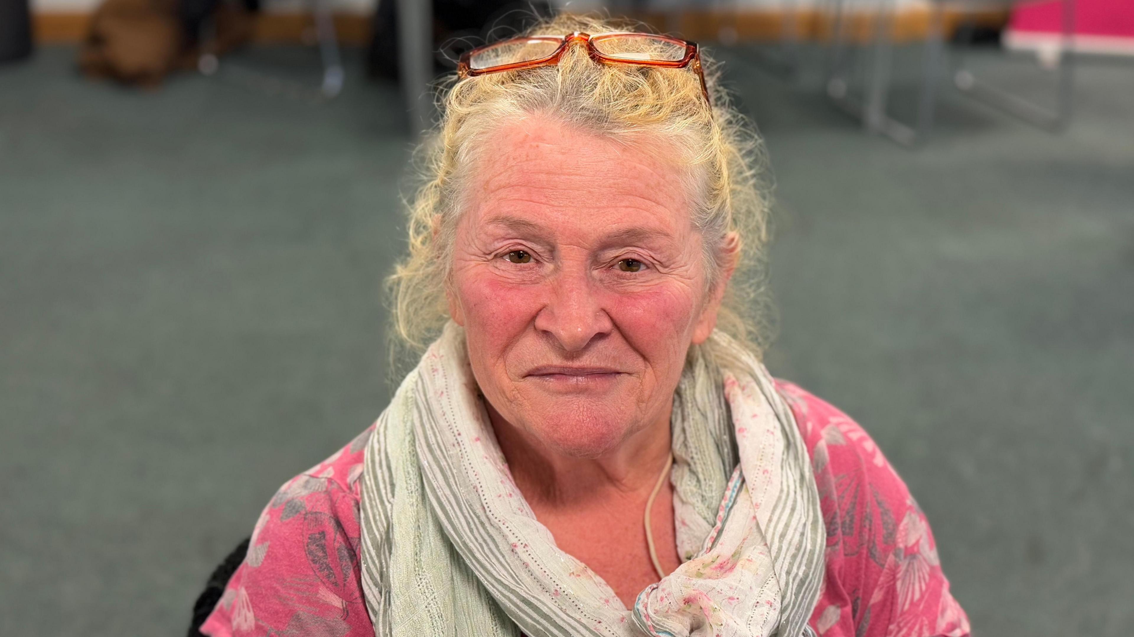 An elderly woman wearing a scarf, pink top and glasses on her head, smiling into the camera 