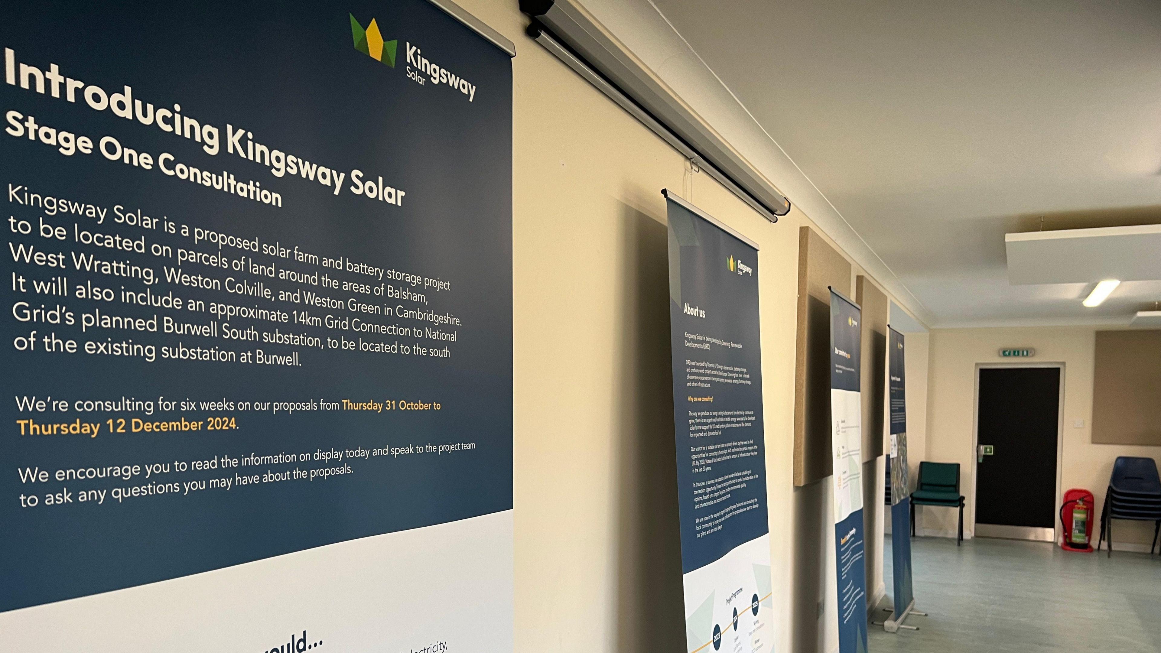 A corridor in a village hall where four presentation posters lean against a wall. The one closest to the camera reads "Introducing Kingsway Solar: Stage One Consultations".  