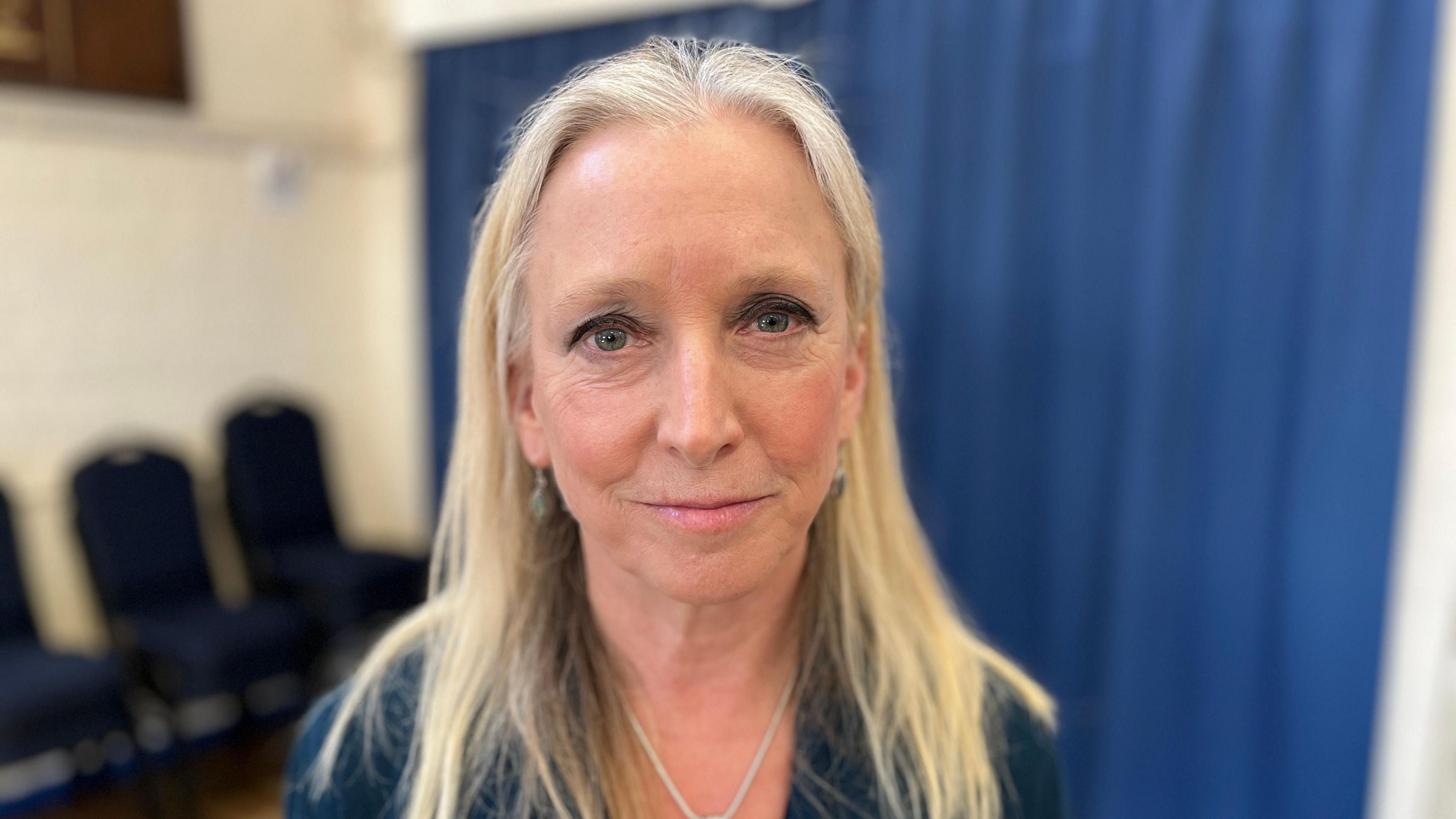 Ros Savage looks into the camera. She stands in front of a dark blue curtain, which is blurred.