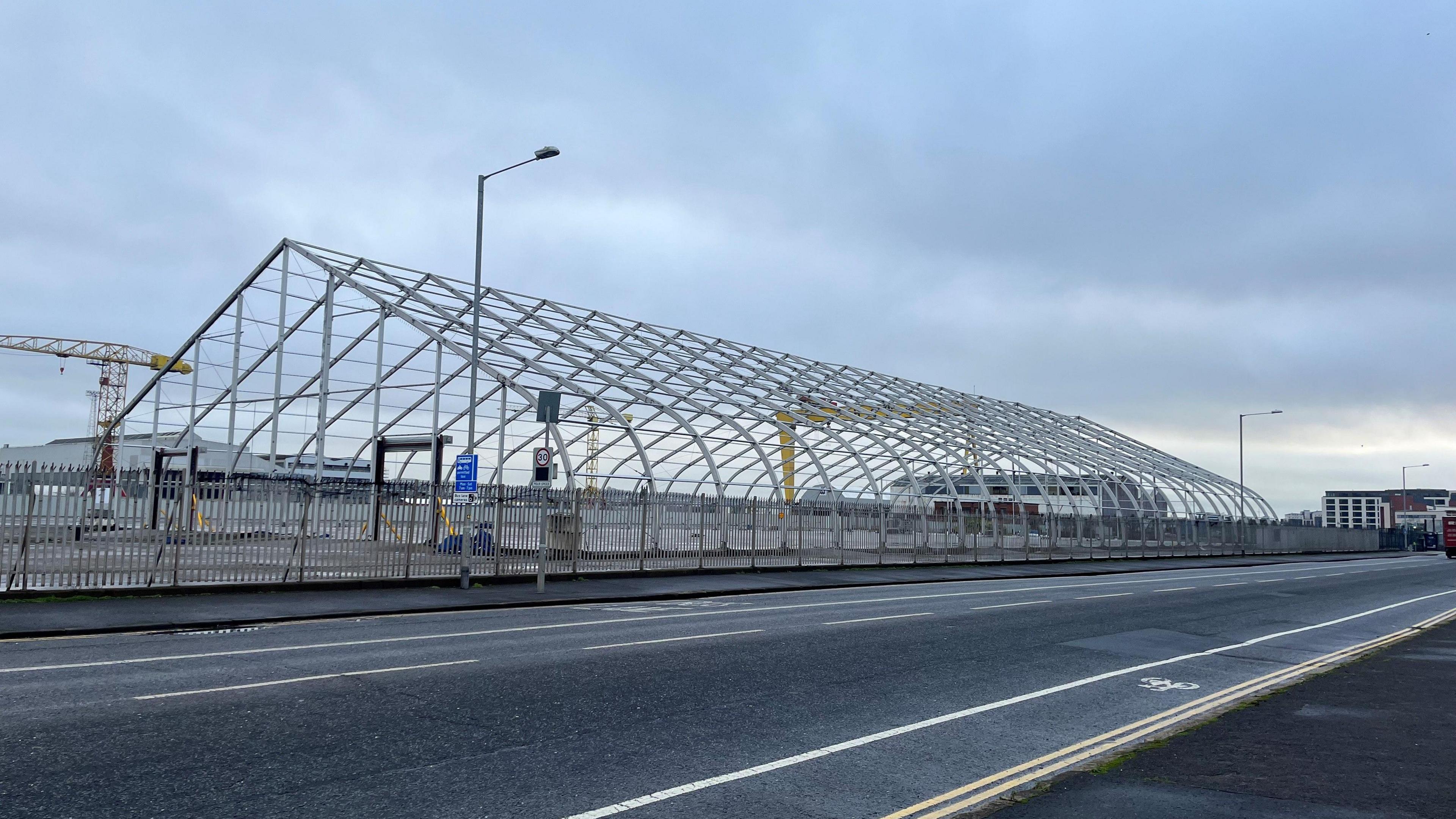 The foundations of the Titanic Exhibition Centre made up of a metal frame. In front of the centre is a road. The sky is grey. The Harland and Wolff yellow crane can be seen in the distance. 