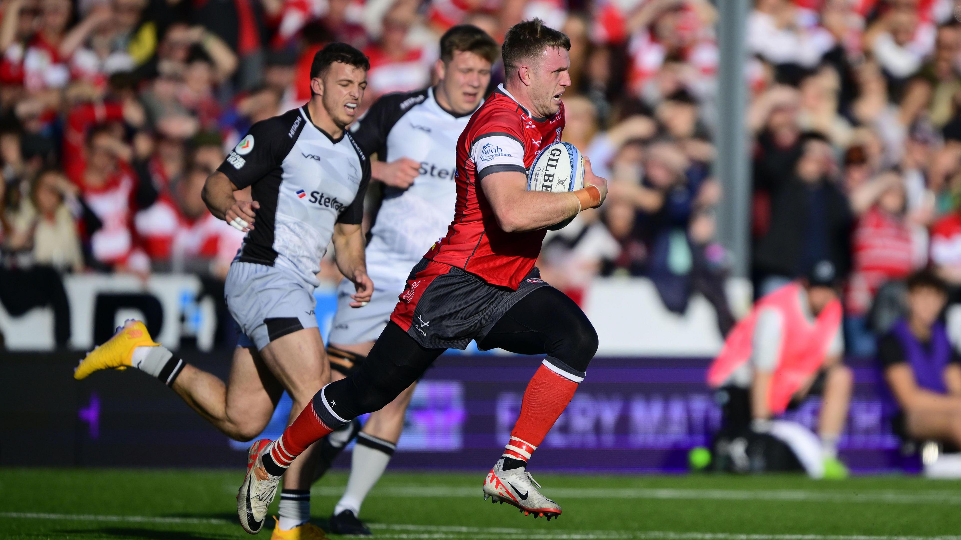 Ollie Thorley runs with a side on profile going to the right of the photo with the ball in his hand during a match for Gloucester against Newcastle, as two Newcastle players chase behind him