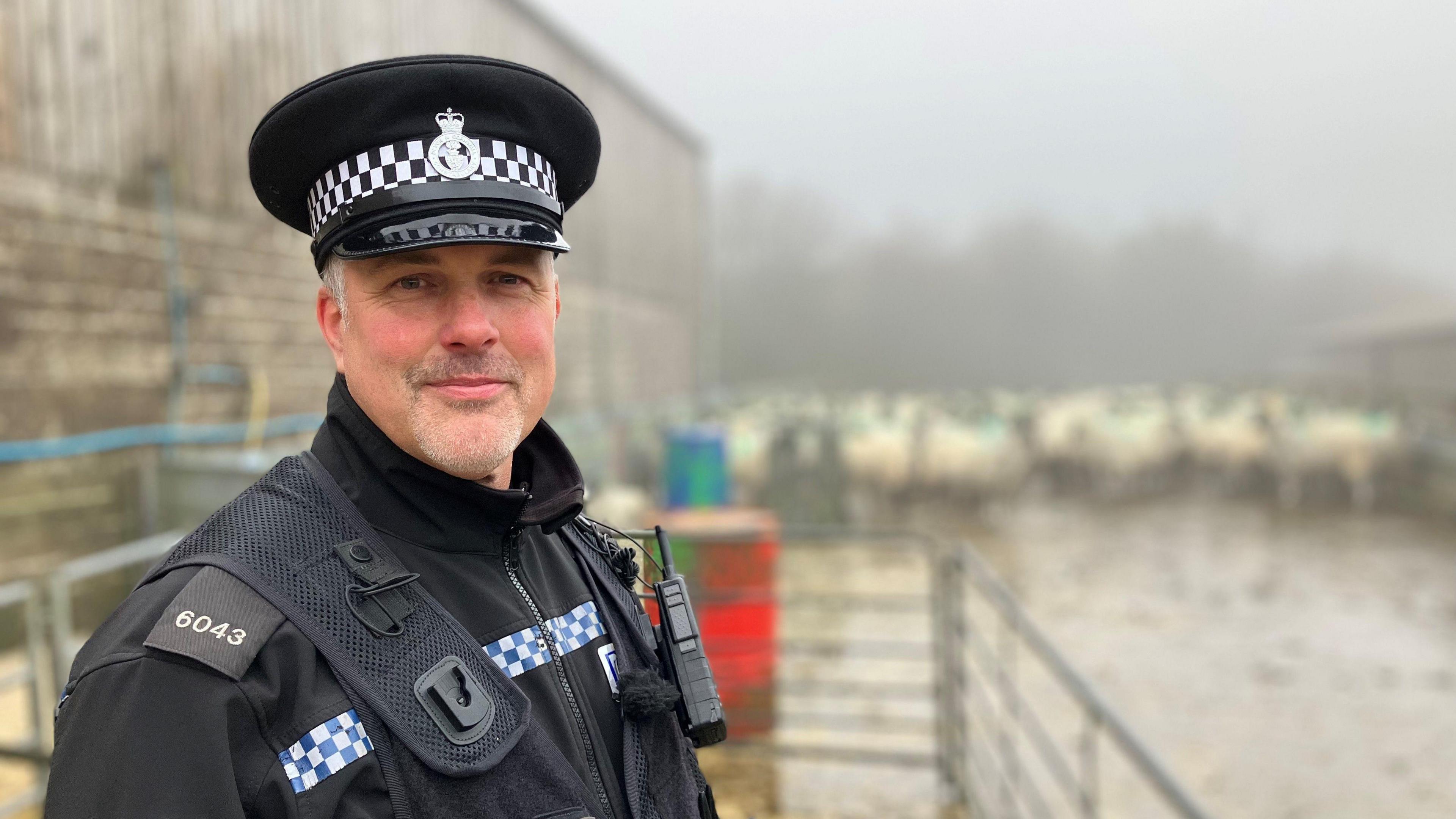 Dressed in a police uniform and hat, PC Julian Fry smiles toward the camera with sheep seen through the fog in the background at a farm in Princetown