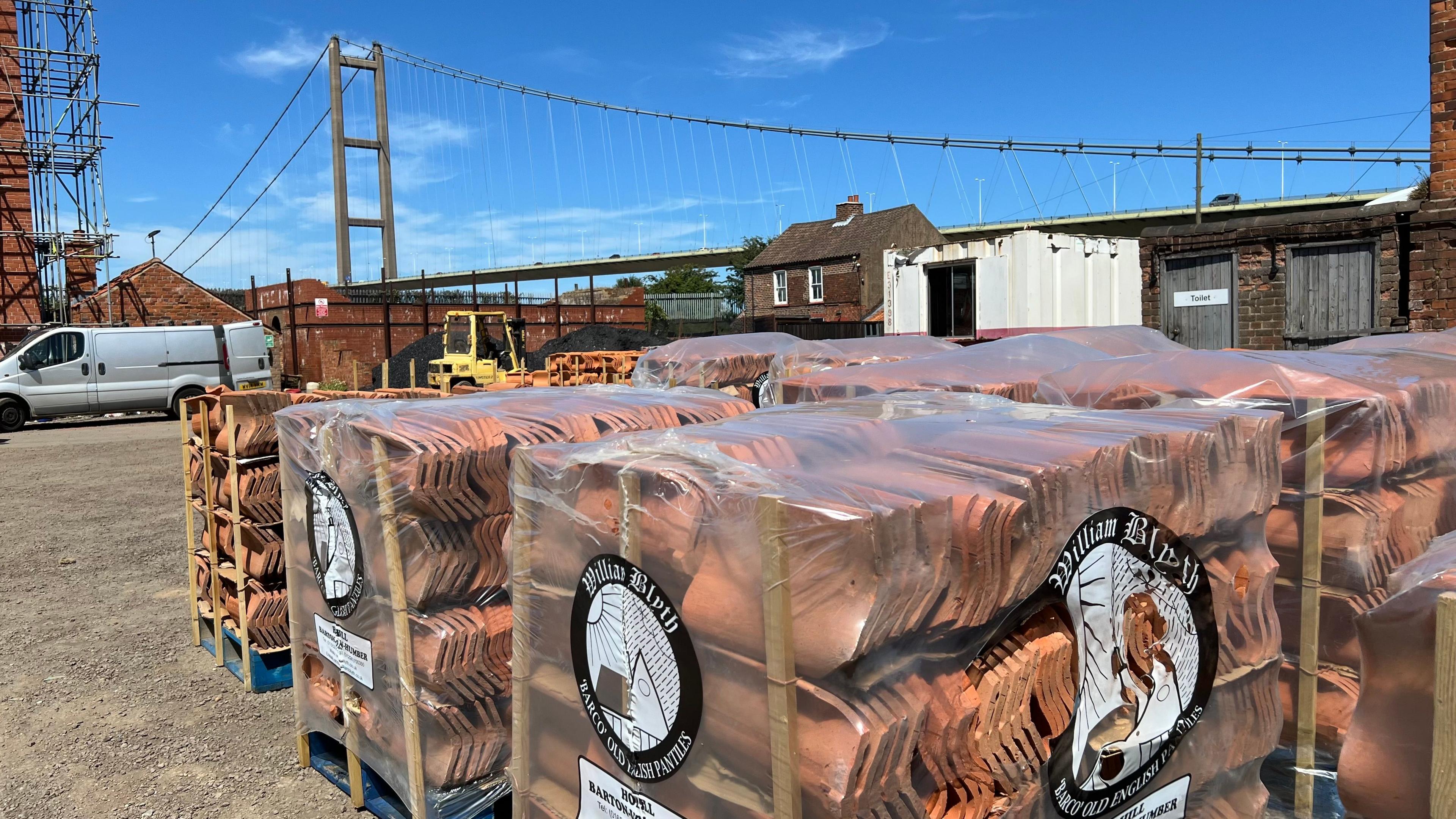 Outdoor view of William Blyth's traditional yard with multiple pallets of terracotta roof tiles wrapped in clear plastic and stacked in the foreground. The packaging displays the William Blyth logo and product labels. Industrial buildings, a white van, and a forklift are visible around the yard. In the background, a large suspension bridge - the Humber Bridge - spans across the scene beneath a bright blue sky with light clouds.