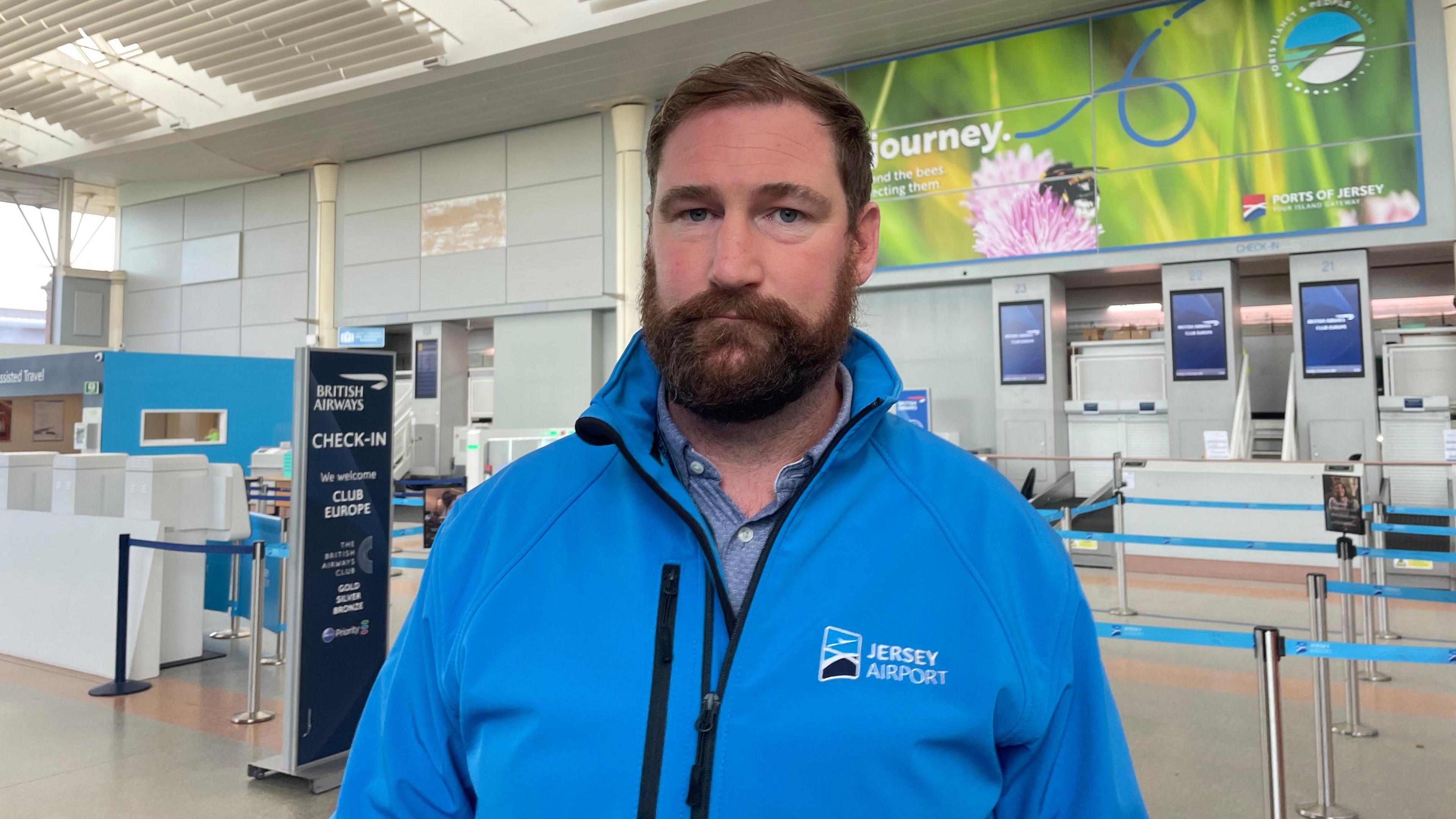 Ashley Maggs - Head of operations Jersey airport wearing a Jersey Airport blue coat standing in front of the British Airways check in area inside the airport.