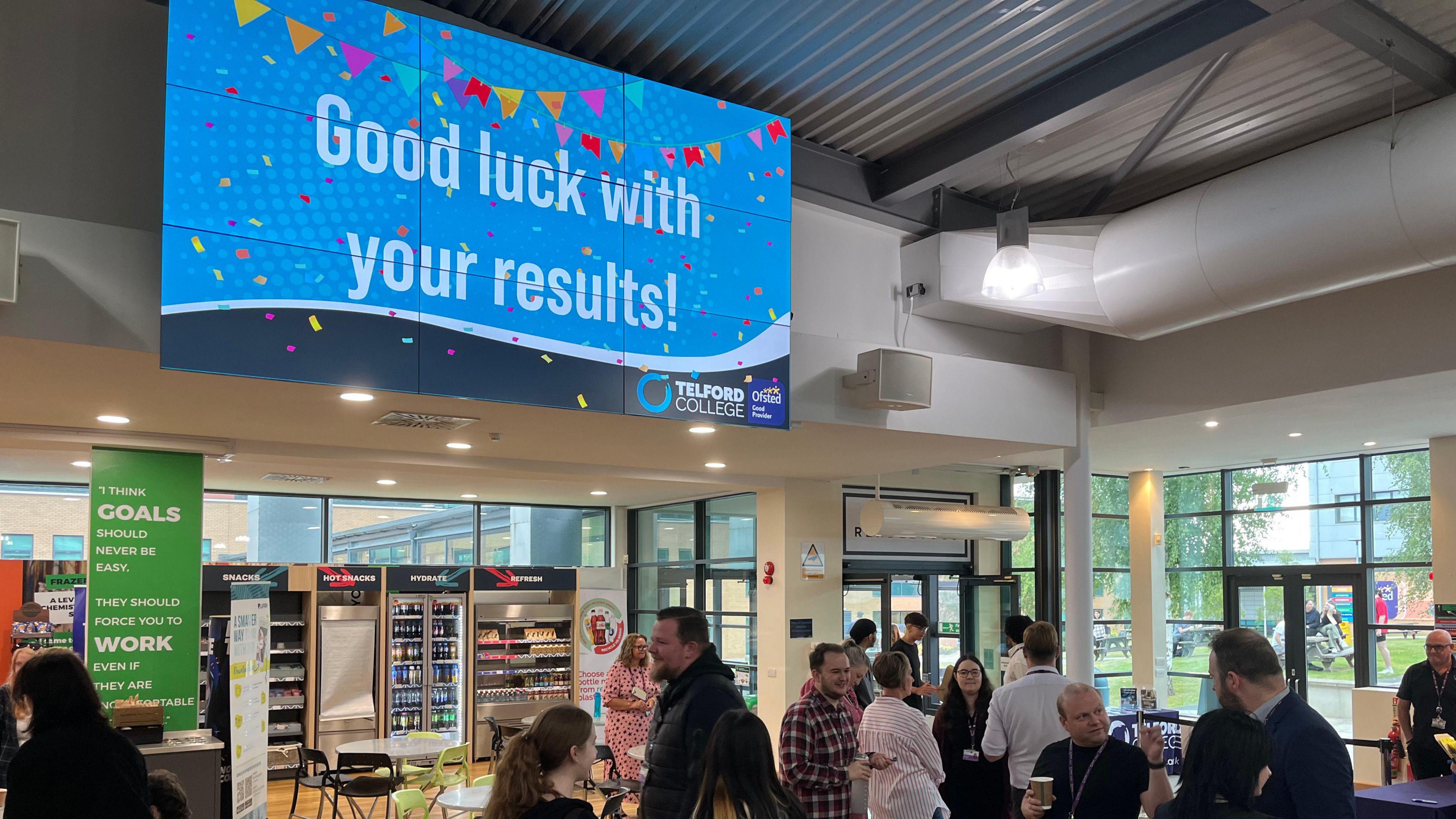 A large television screen is mounted on a wall near the ceiling of a college building. The screen is blue with colourful banners, and reads "good luck with your results!". There are people walking below it.