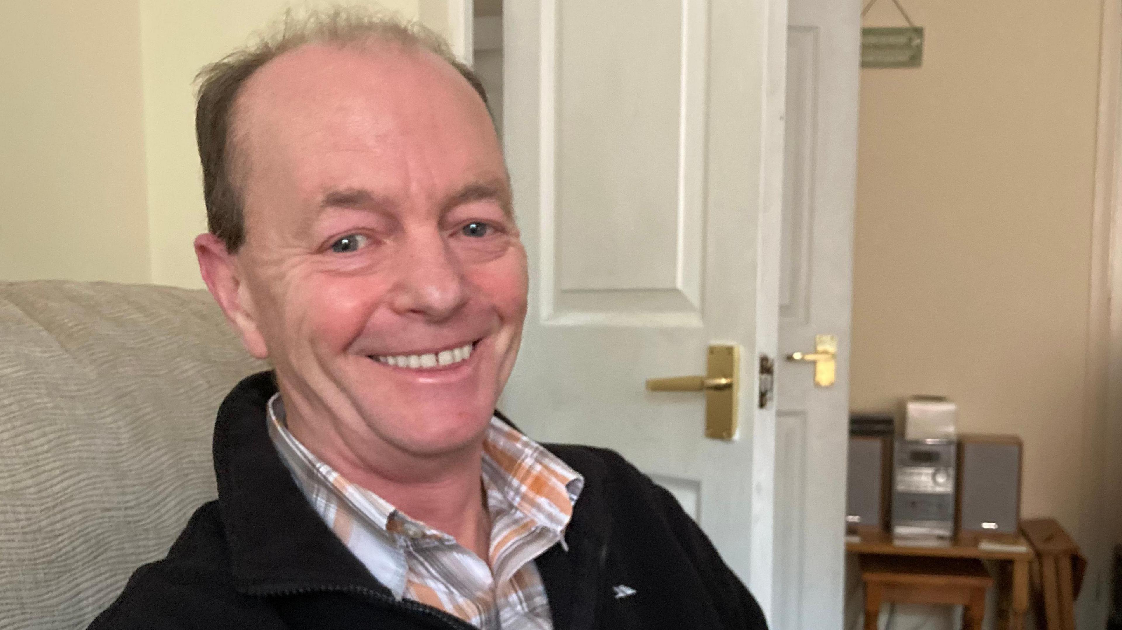 Mark Abel at home, sitting on a sofa, wearing a shirt and black jumper. He is smiling at the camera and looking happy and excited.