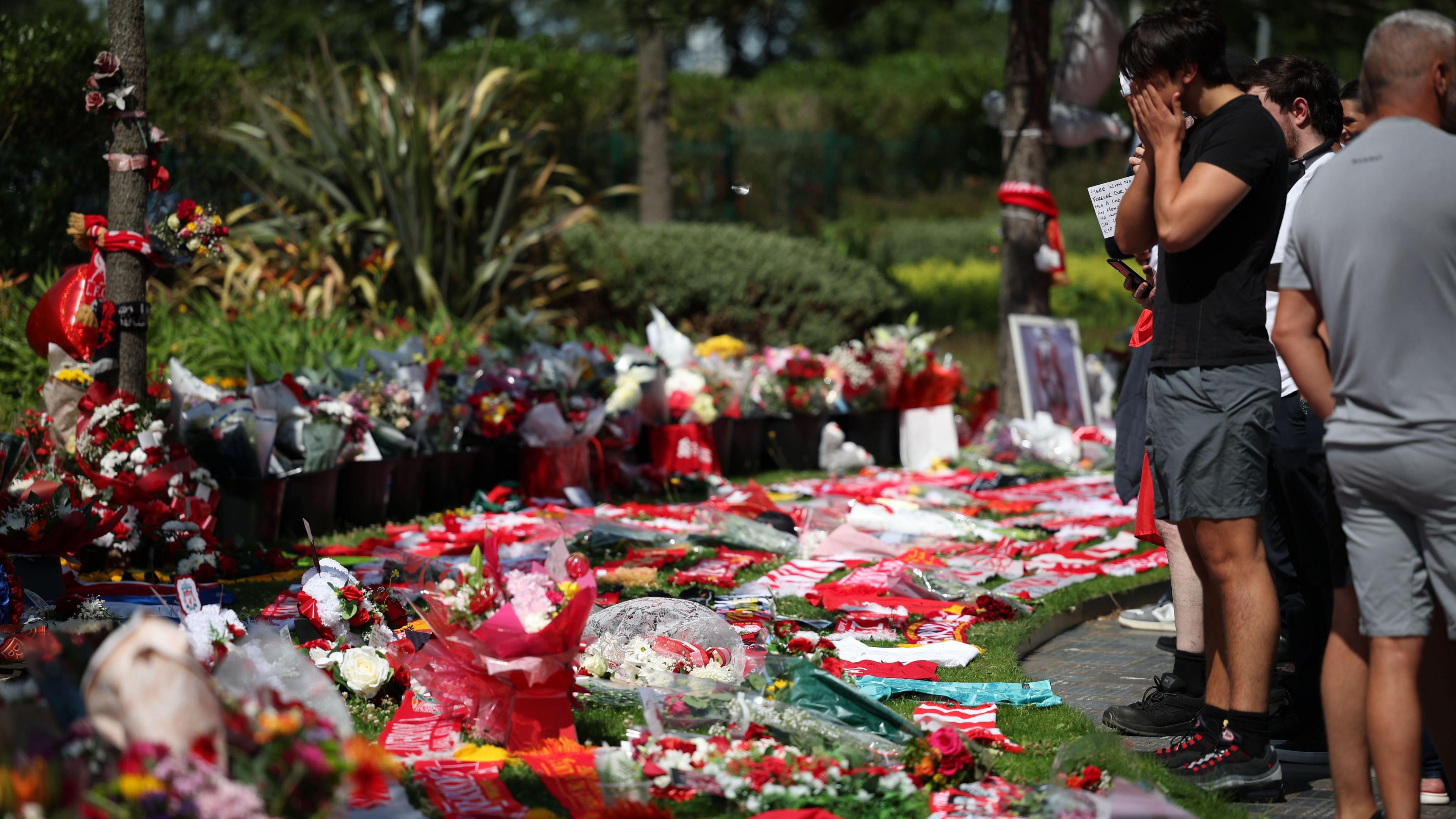 A crowd of people, including a man with his face in his hands, stand on the edge of a grassy area blanketed in flowers, football shirts, scarves and balloons