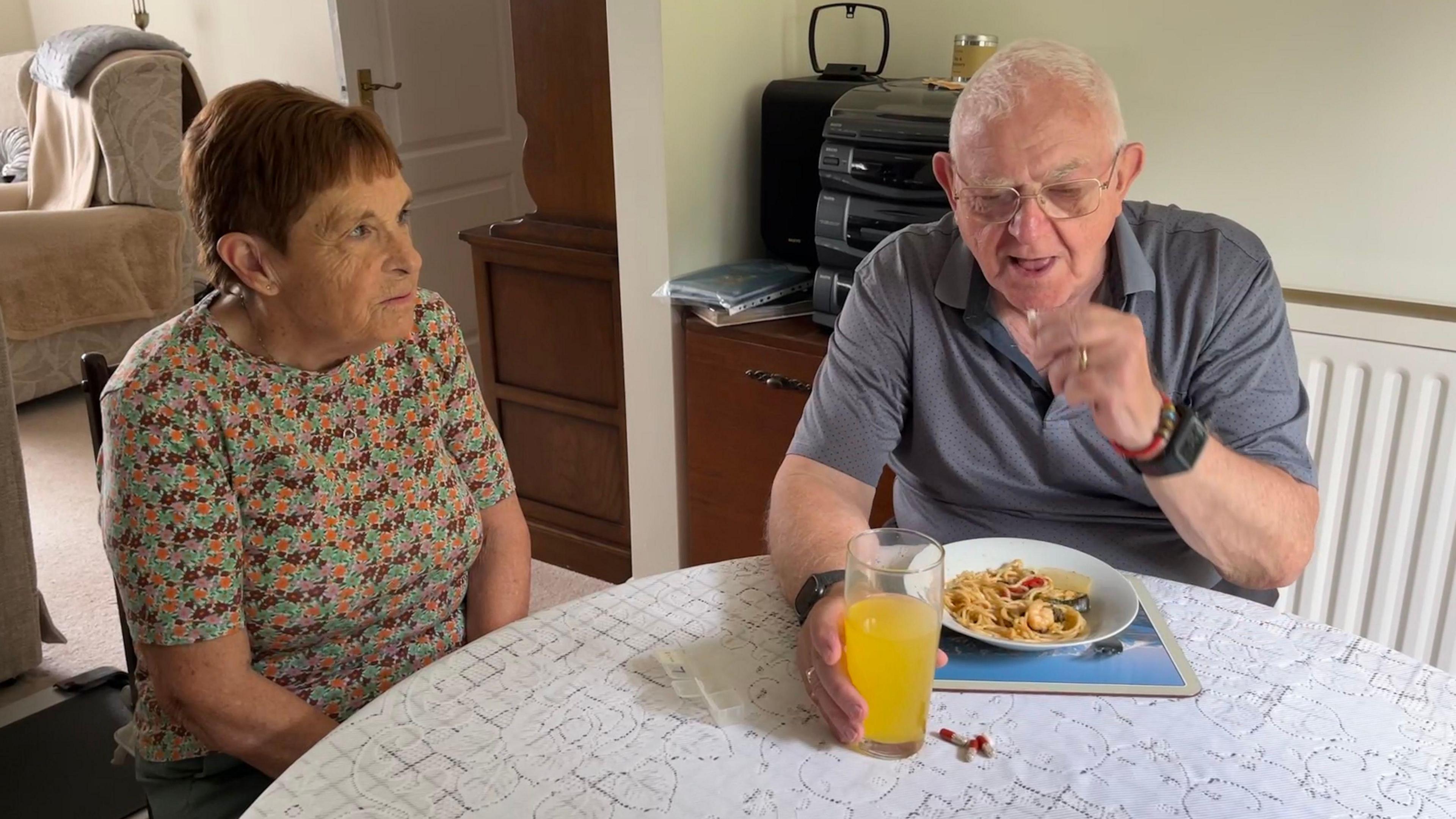 Mr Allen sitting at a table with a bowl of pasta in front of him, taking a Creon capsule with a glass of orange squash