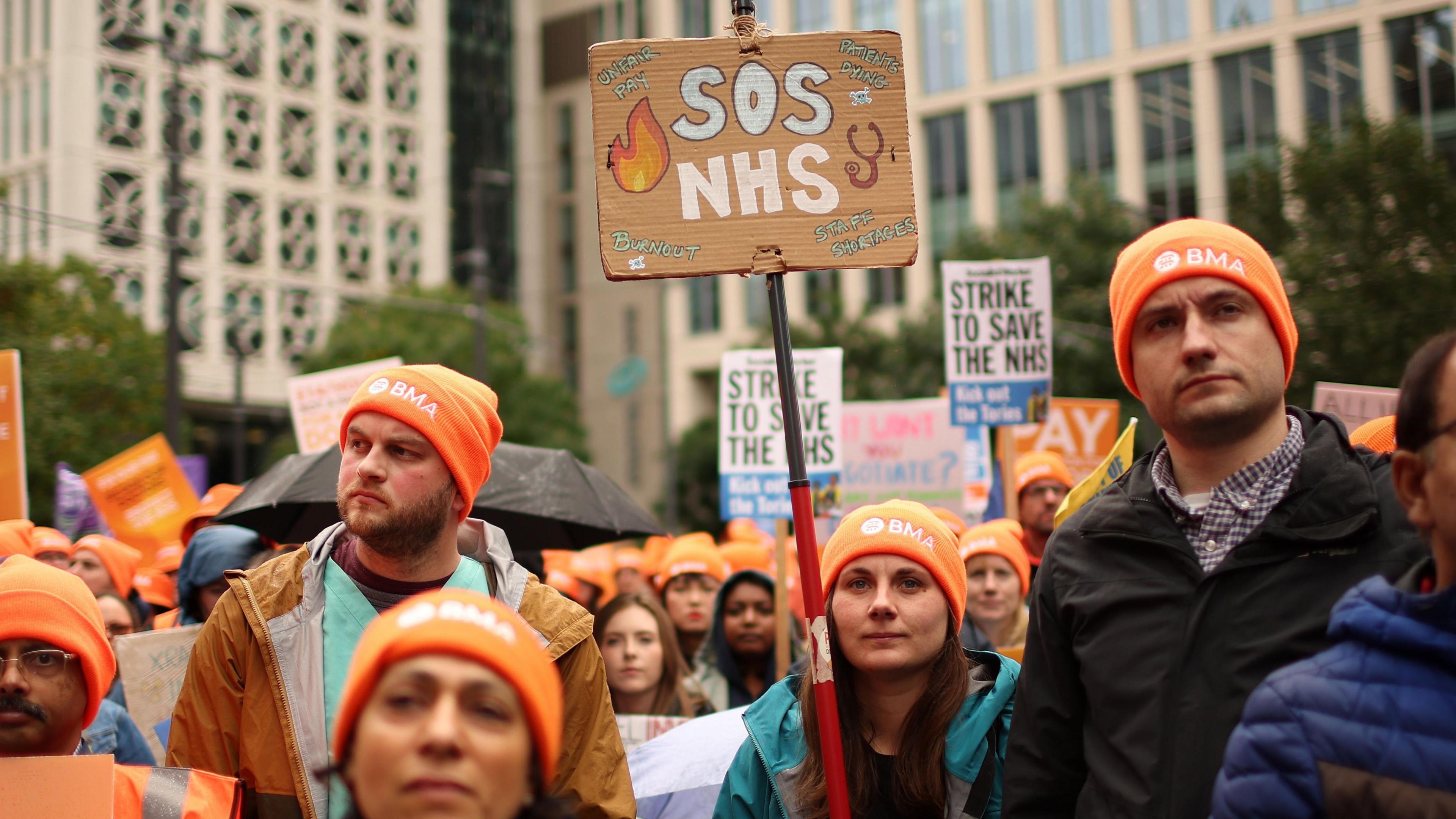 People in orange hats with BMA written on them gather in Manchester. A woman in the middle is holding a sign that says 'SOS NHS'