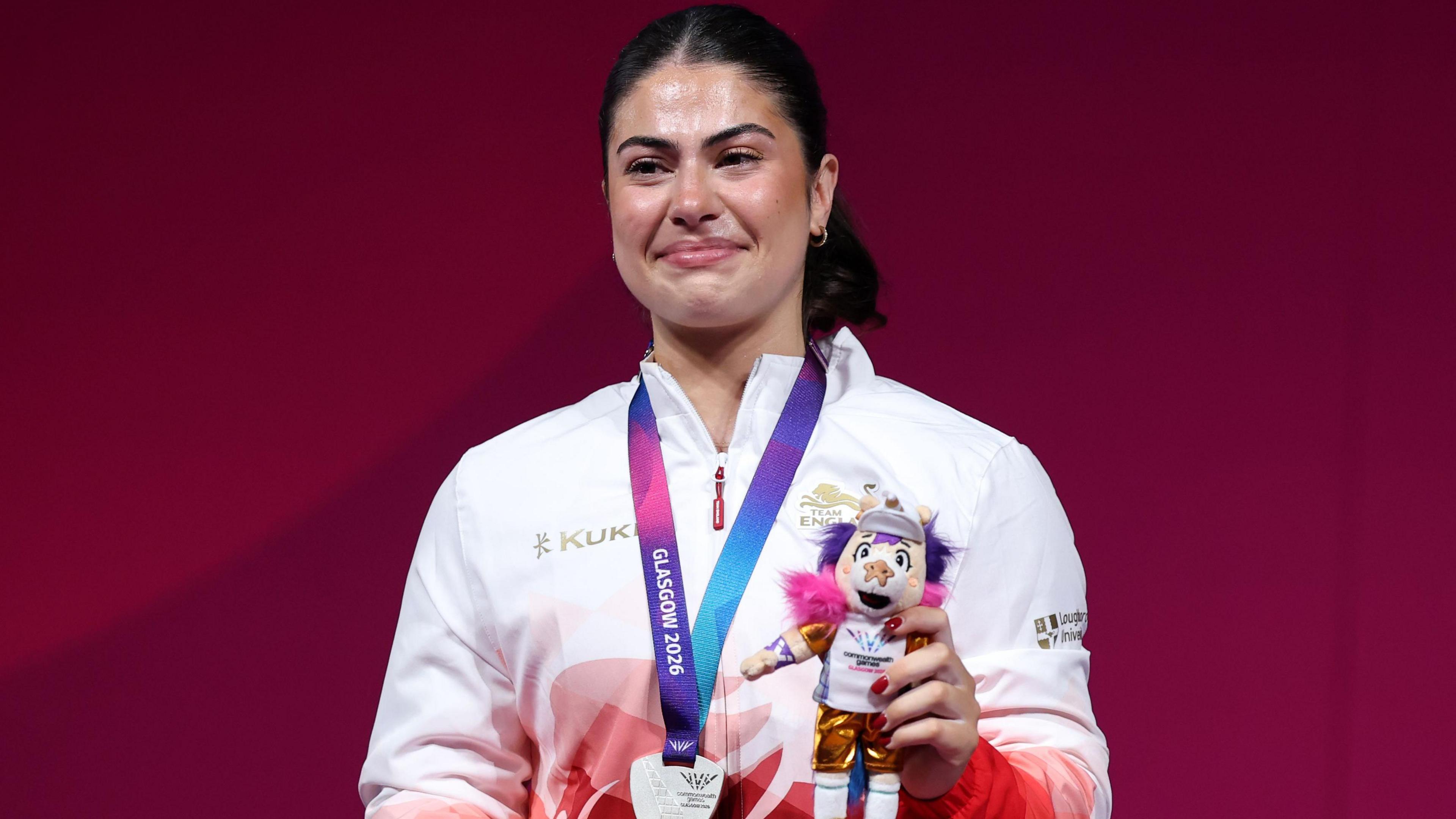 Isabella Brown, wearing England's white and red jacket, holds up a mascot after winning silver in the 77kg women's weightlifting at the Commonwealth Games. Her silver medal is around her neck on a multicoloured pink, blue and purple ribbon.