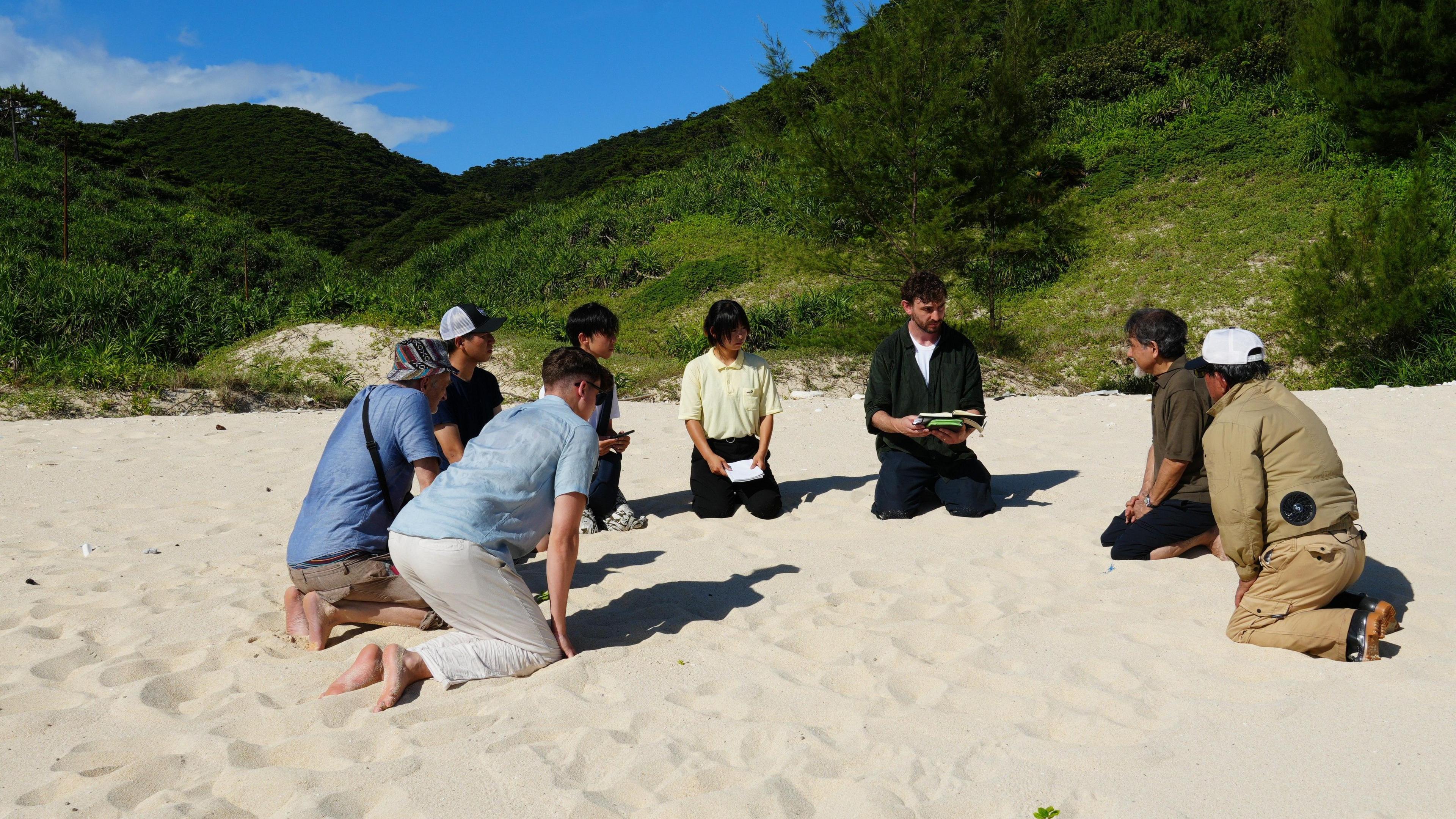 A group of 8 people are kneeling in the sand on a beach on Aka island in Japan. Behind them is a steep cliff covered in green vegetation