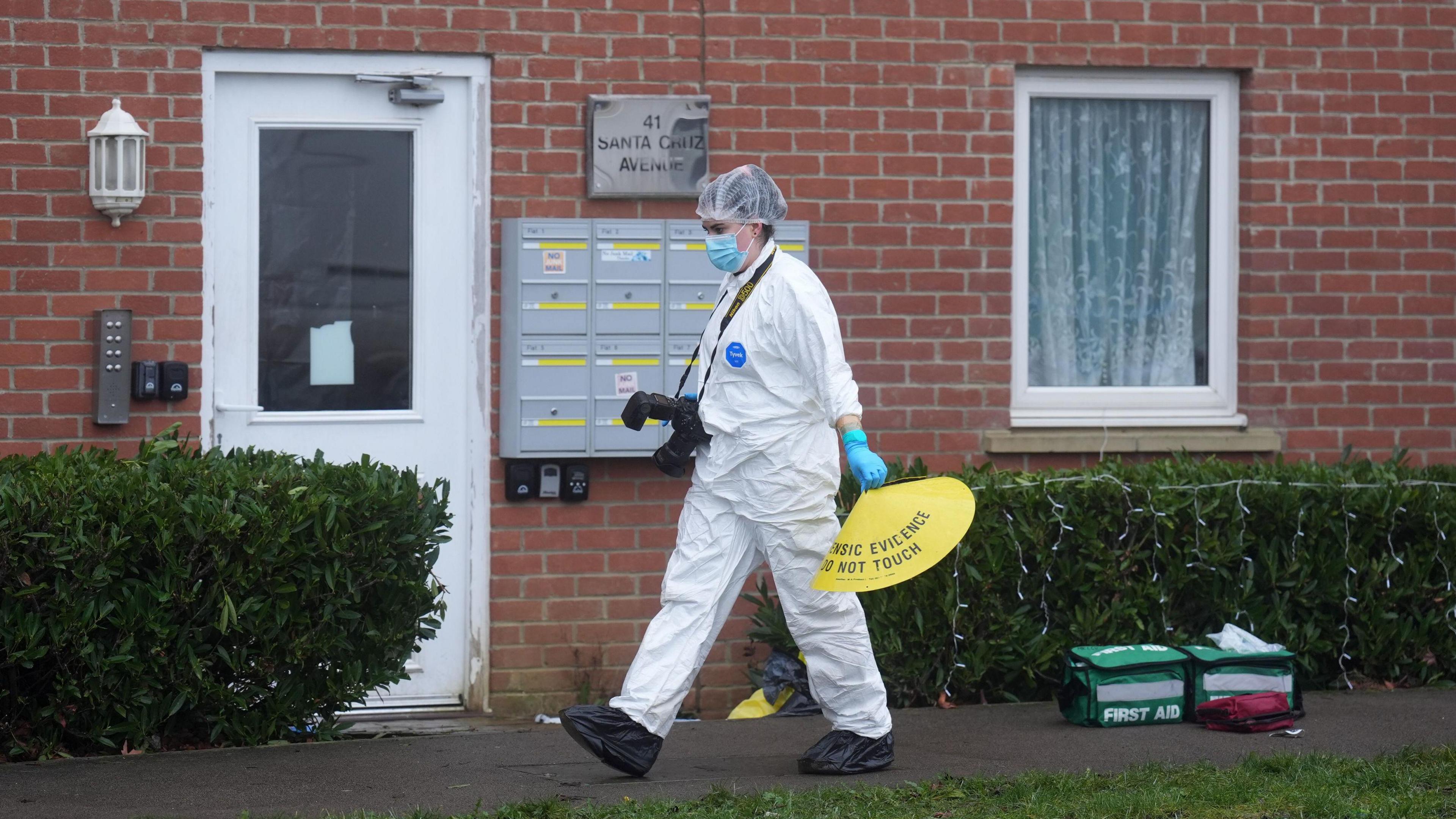 A forensics officer, in a white suit, hair net and face mask, is walking from right to left outside terraced housing. They have a camera and are holding a yellow cone. There are two green first aid bags on the pavement outside the houses.