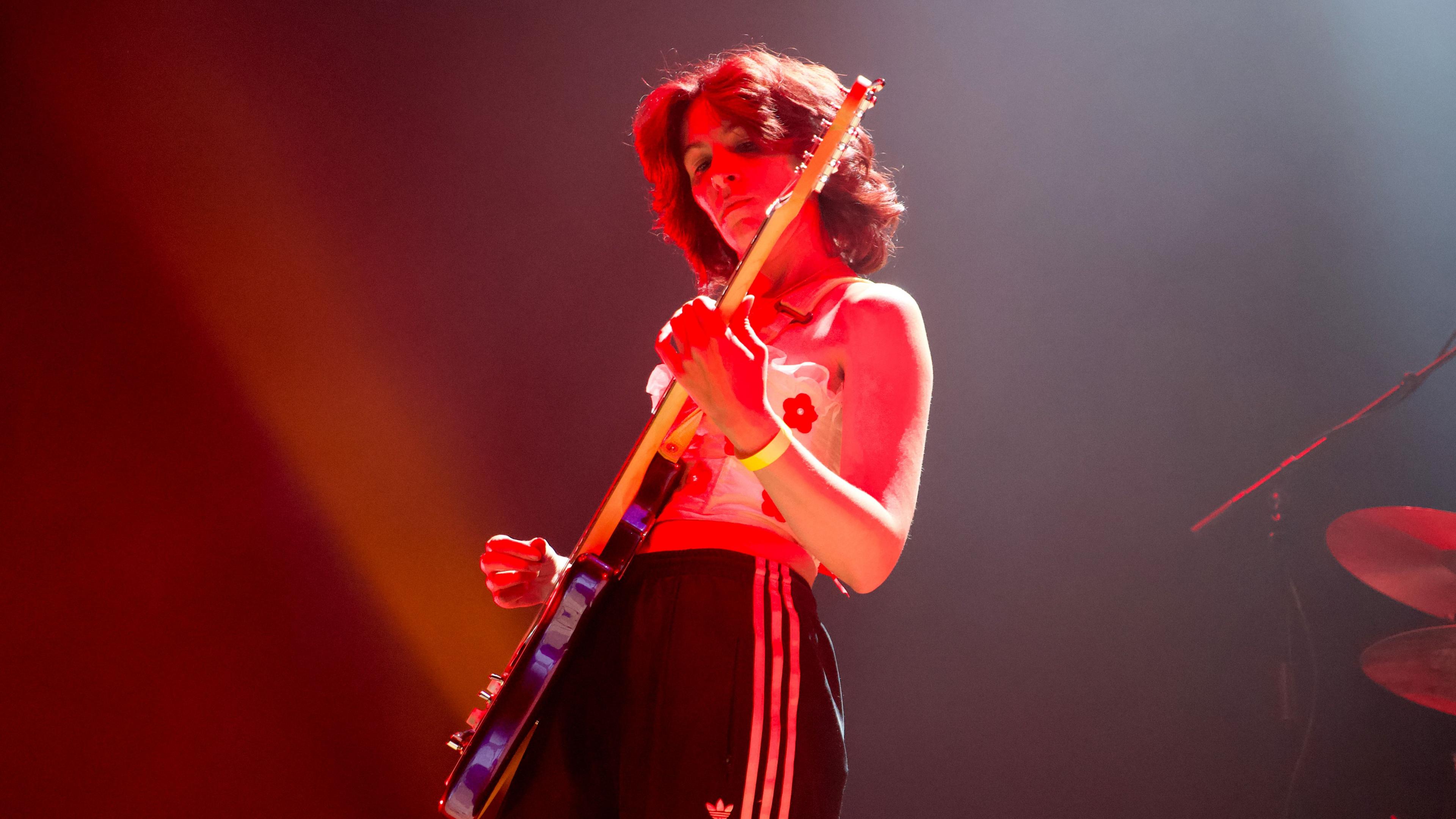 A woman in a white top and black tracksuit bottoms playing guitar